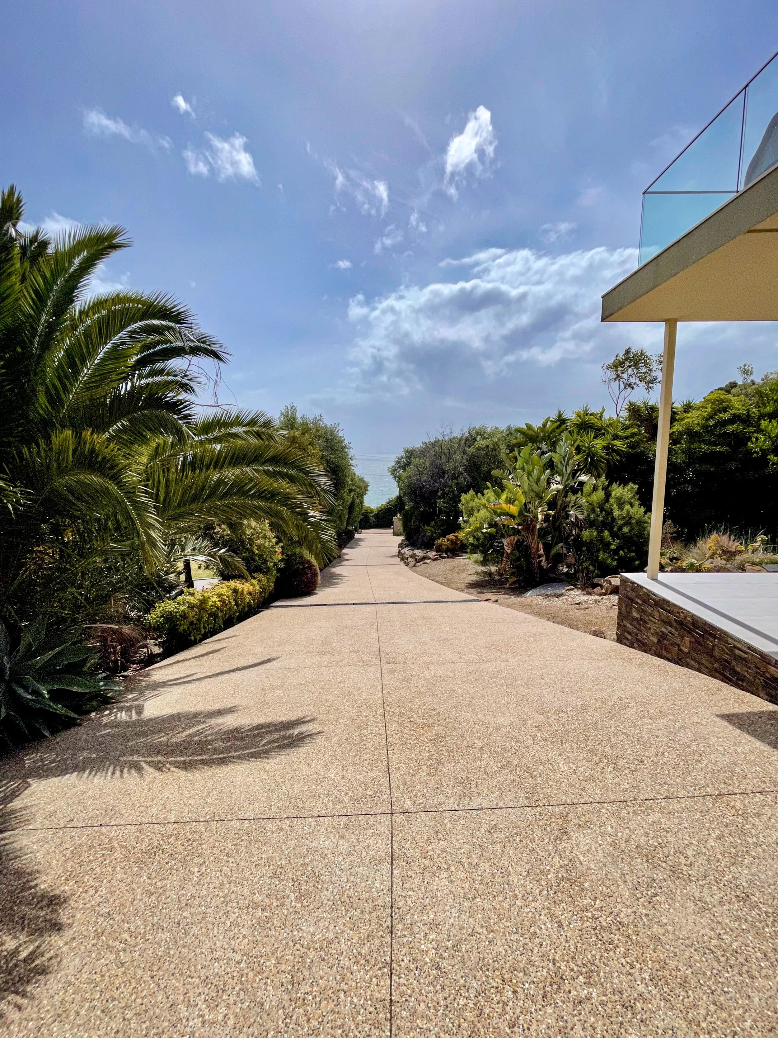 A paved walkway in a lush garden with various green plants and trees on either side, leading towards a distant view of the ocean under a partly cloudy sky.
