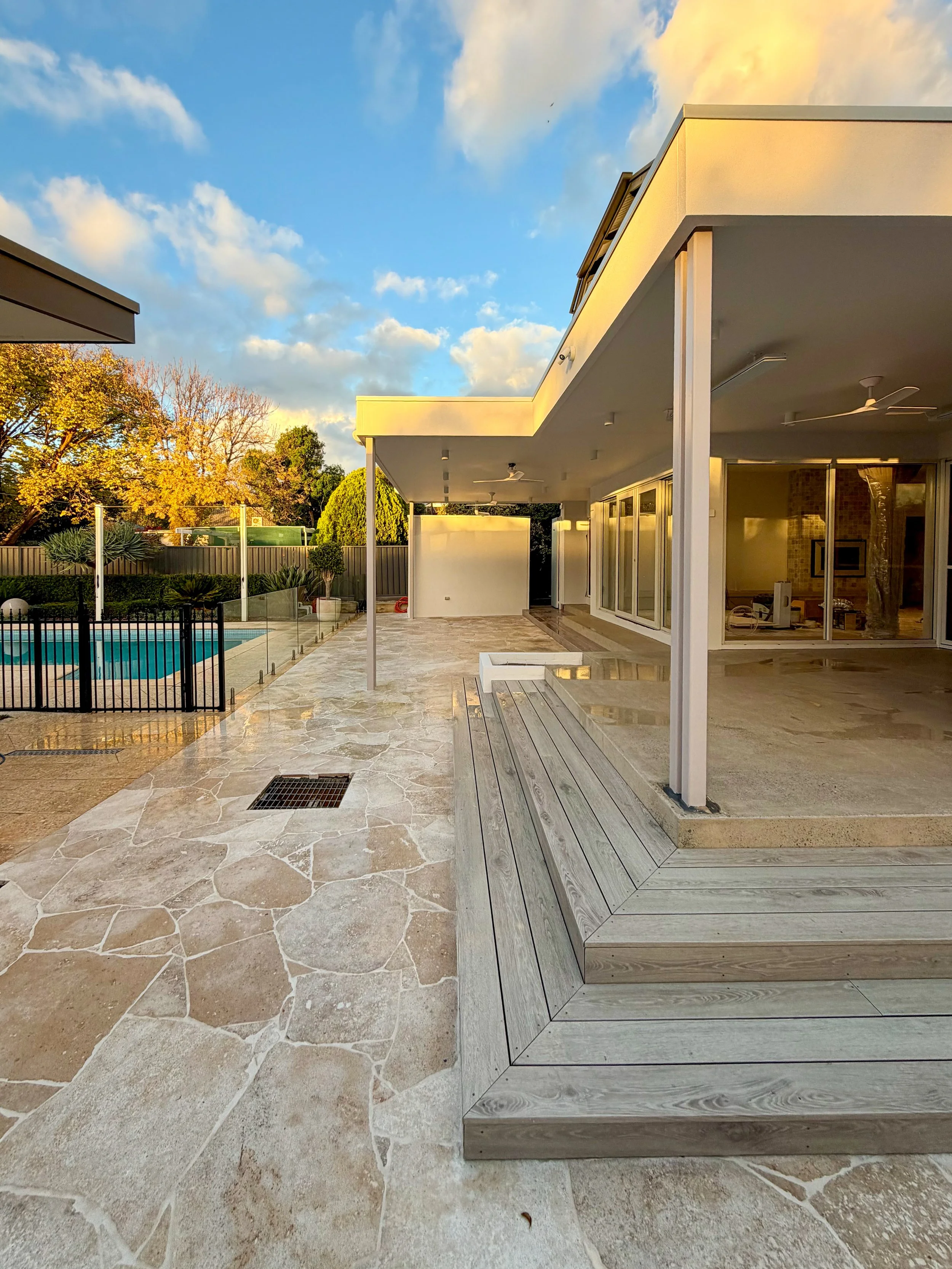 A modern house with a large covered outdoor patio, glass sliding doors, a swimming pool, and trees in the background under a blue sky with clouds.