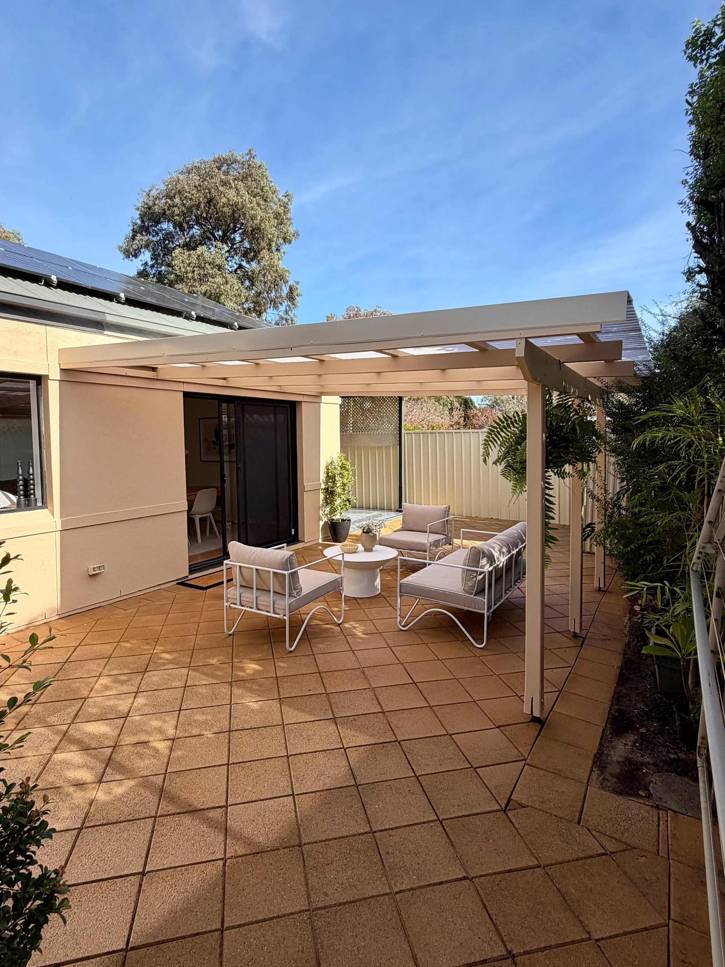 A backyard patio with terracotta tiled flooring, white outdoor furniture including a table and chairs, a beige structured pergola, and surrounding greenery including trees and plants under a blue sky.