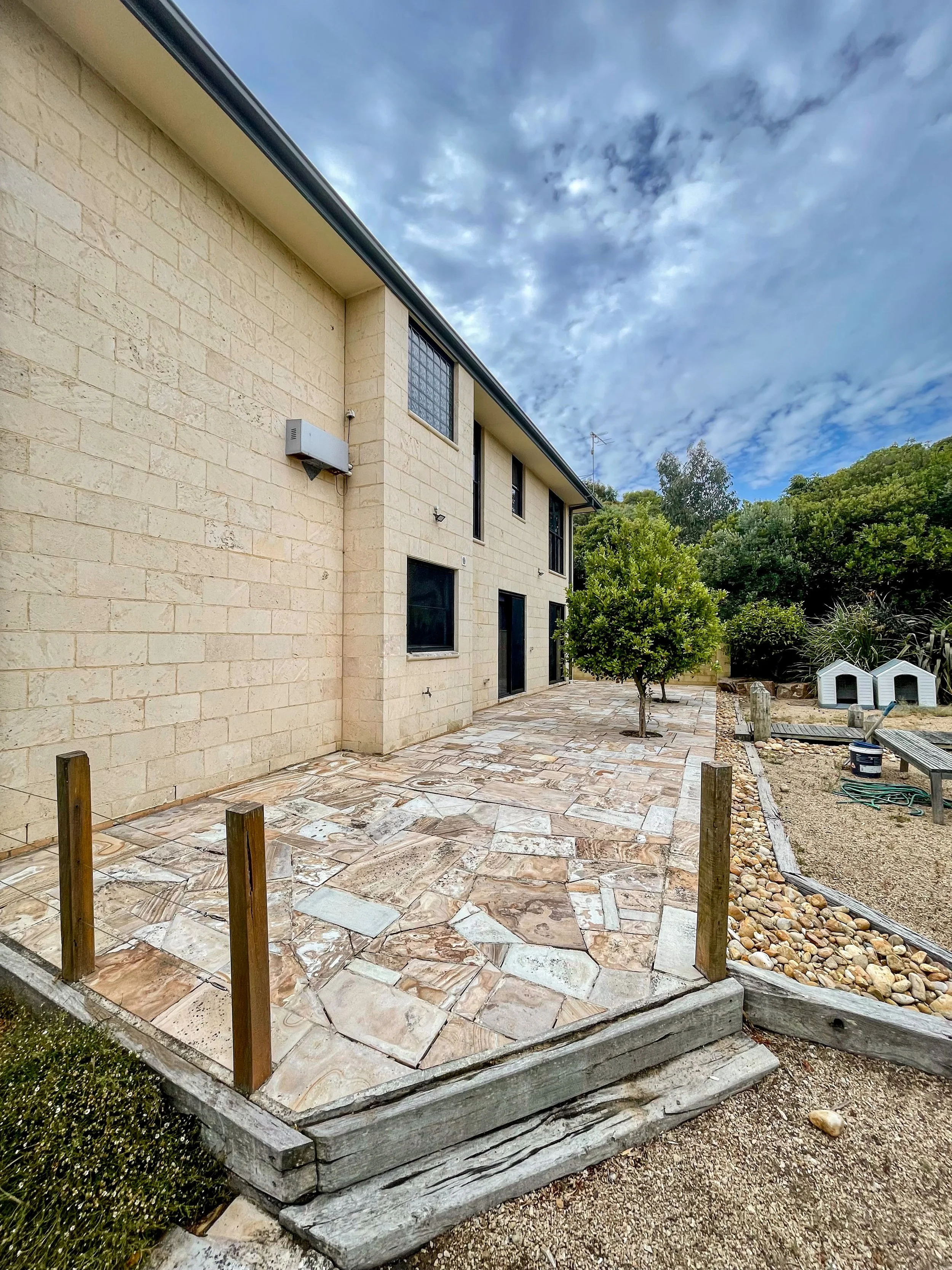 Backyard patio with uneven stone tiles, small trees, and a beige house exterior with three windows. Early stage of outdoor renovation with wooden posts marking the area. Clouds in the sky.