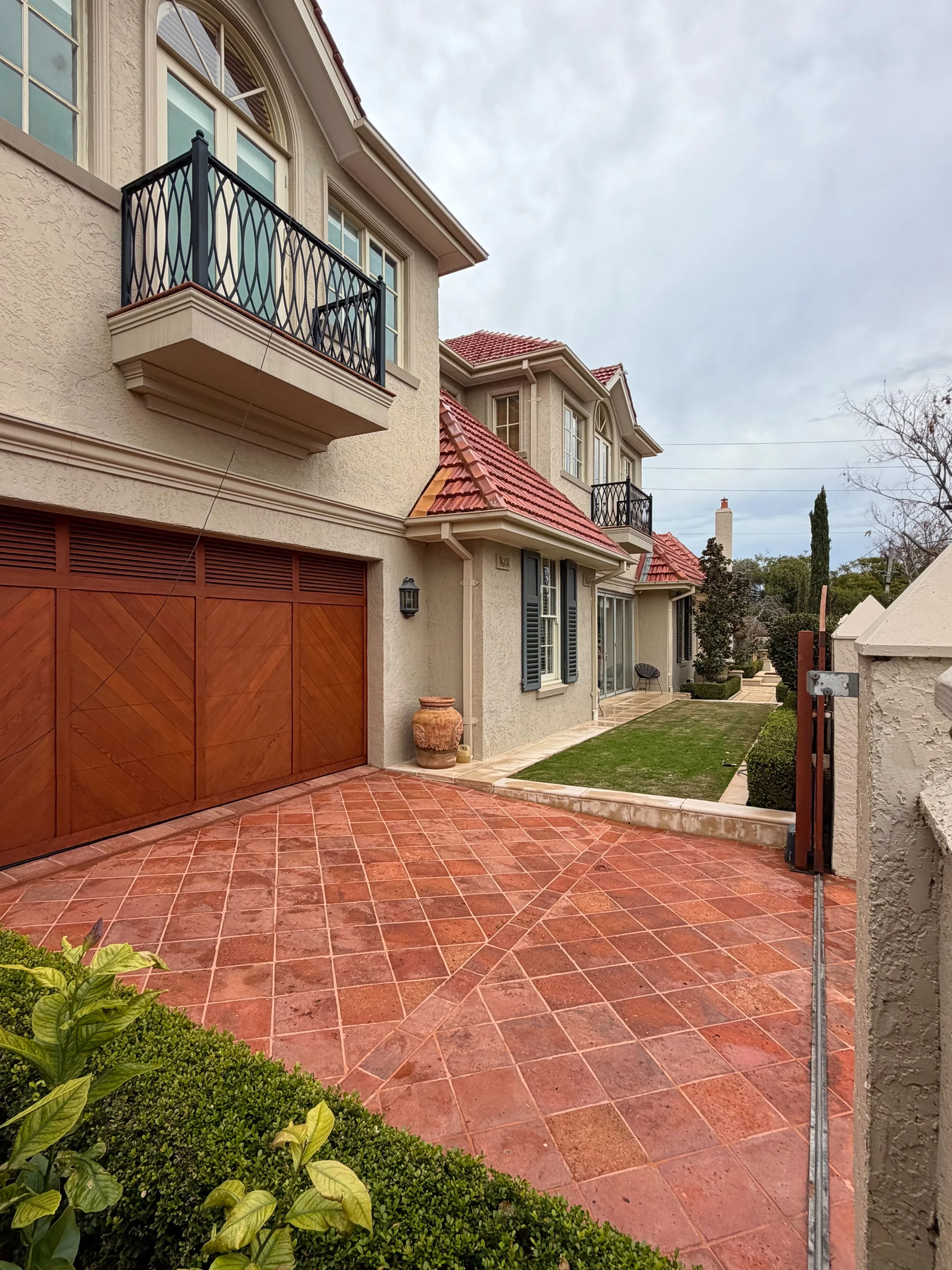 View of a house with a red tile roof, beige stucco walls, a wooden garage door, and a small balcony with black metal railings, a patio area with red tiles, and some shrubbery in the garden.