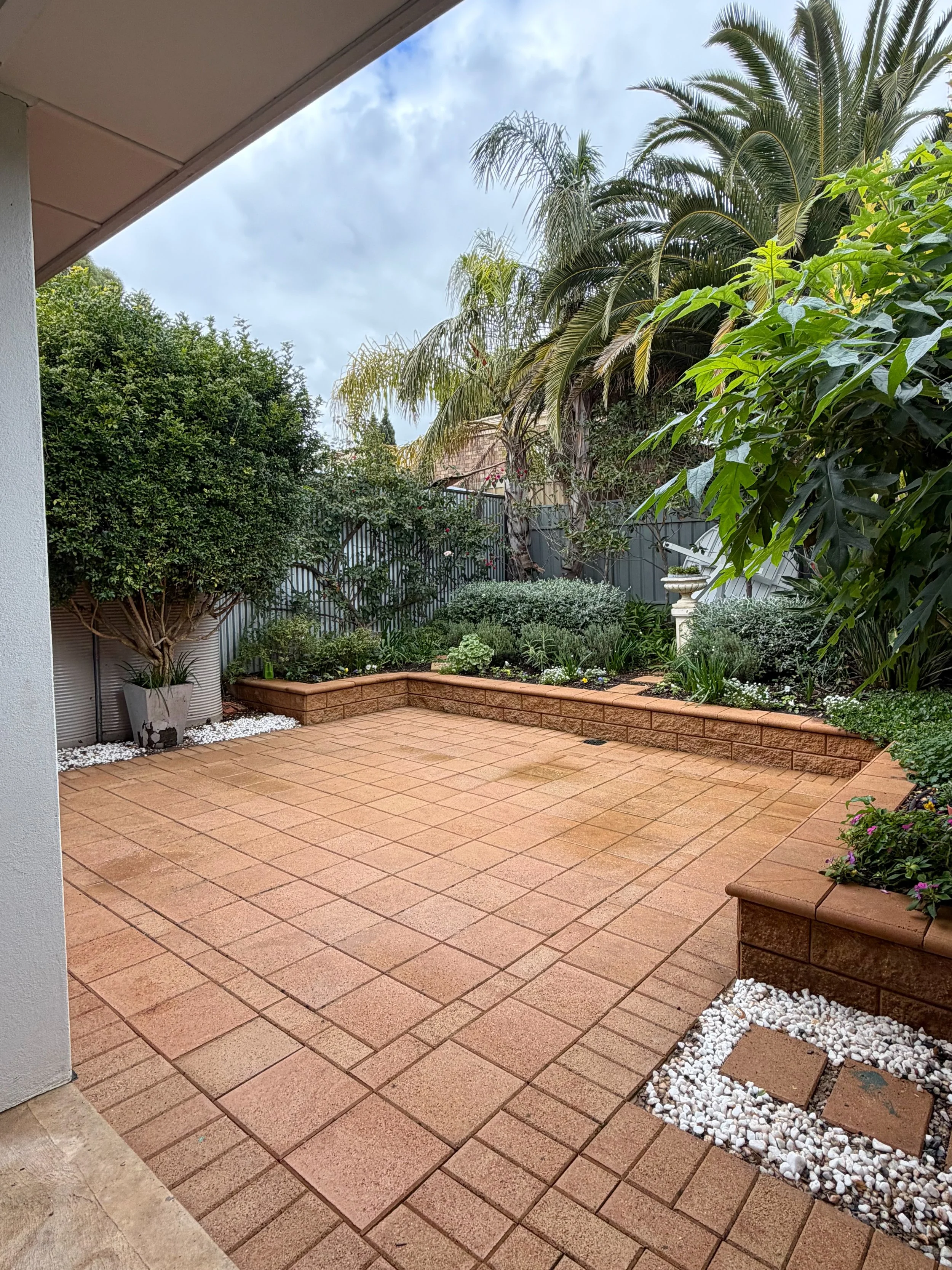 A backyard patio with brown brick pavers, a garden bed with plants and small flowers, a birdbath, and a lush hedge and palm trees in the background. The sky is partly cloudy.