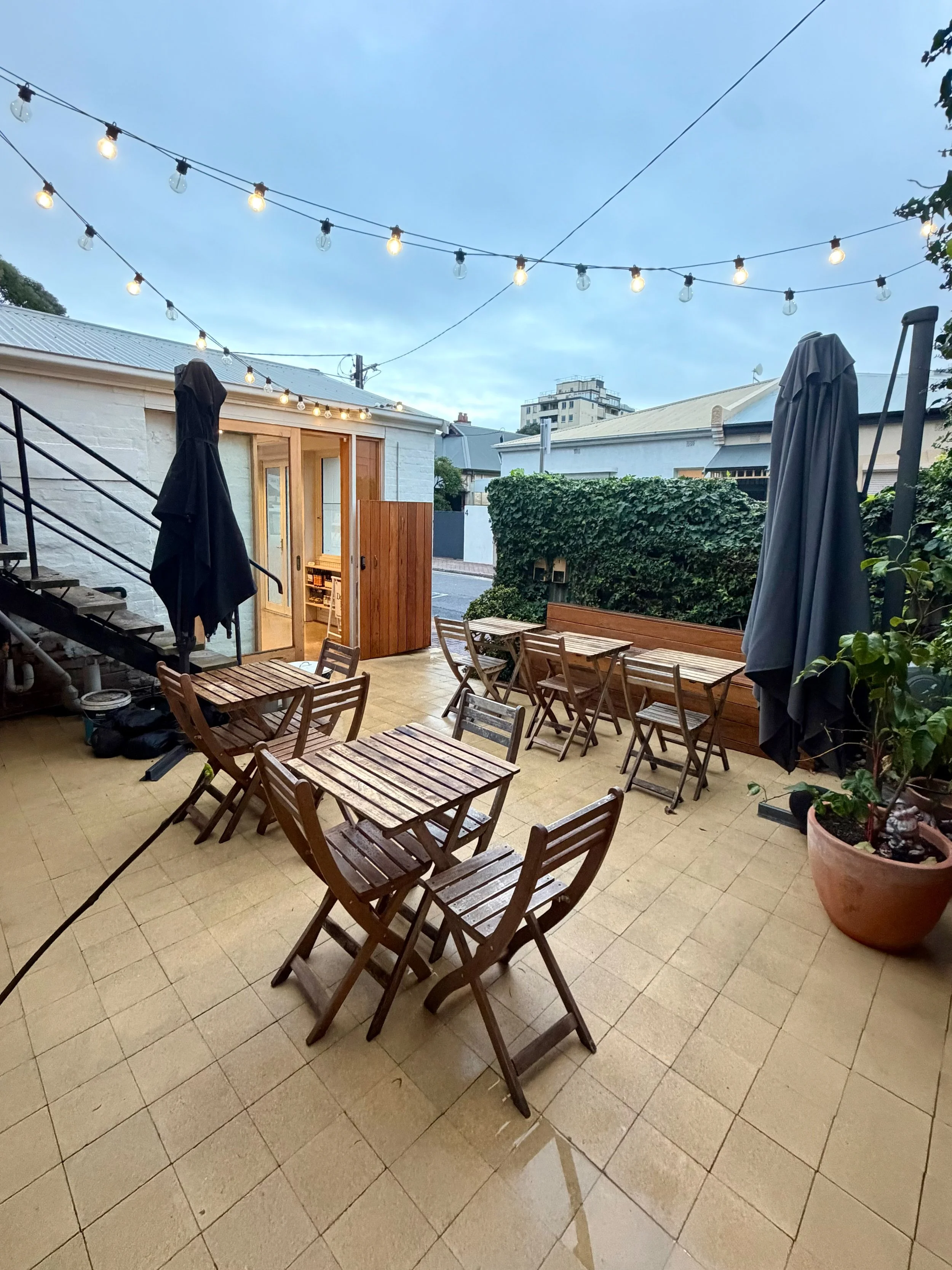 An outdoor patio area decorated with string lights, wooden tables and chairs, potted plants, and umbrellas. There is a small building with a door in the background.