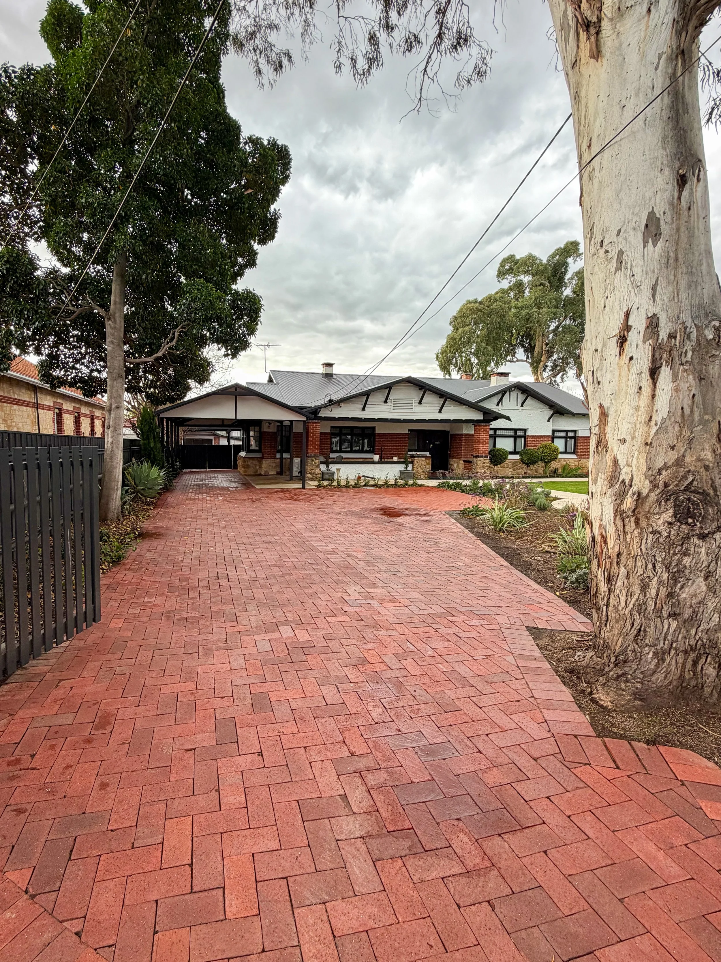 A house with a brick driveway and sidewalk, surrounded by trees and bushes under a cloudy sky.