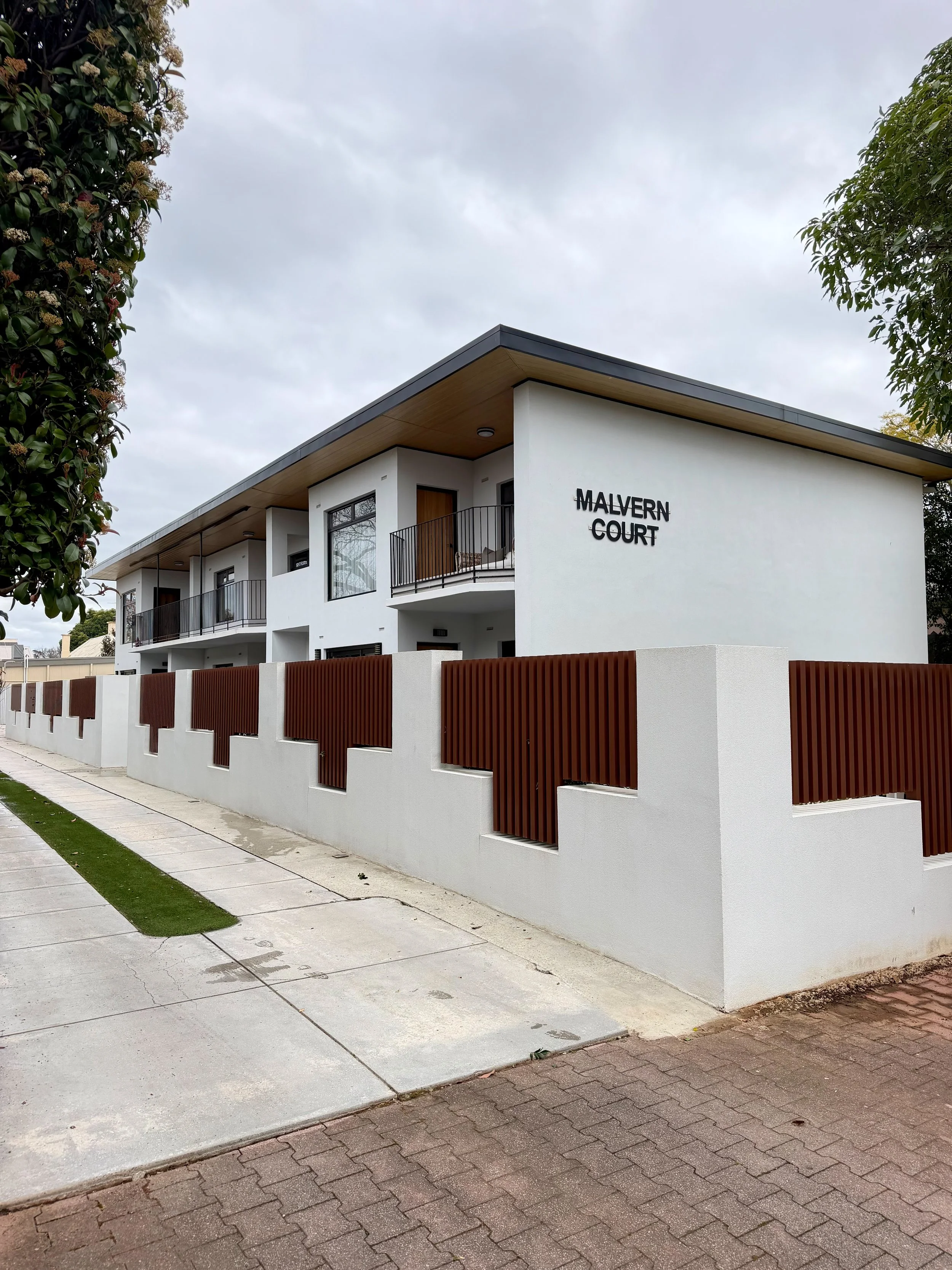 Modern white residential building with balconies and black railings, surrounded by a white wall with brown wooden accents. The sign on the building reads 'Malvern Court'. There is a sidewalk in front of the building with a strip of grass and neighbor