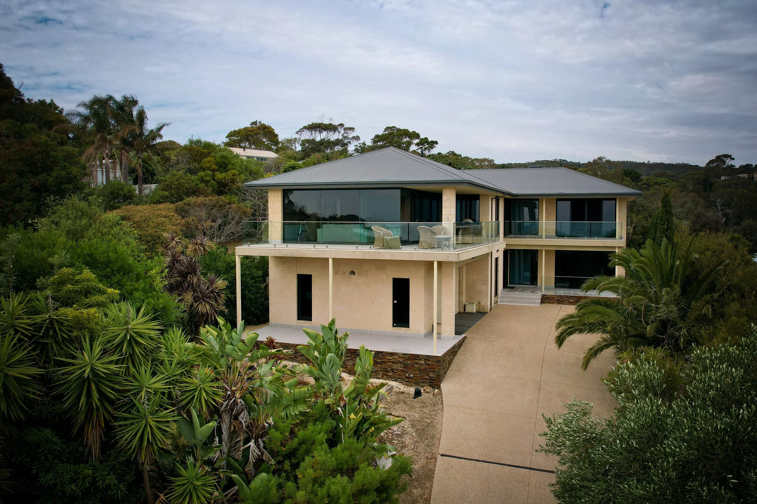 Modern house with glass railing balcony surrounded by lush green trees and bushes, on a cloudy day.