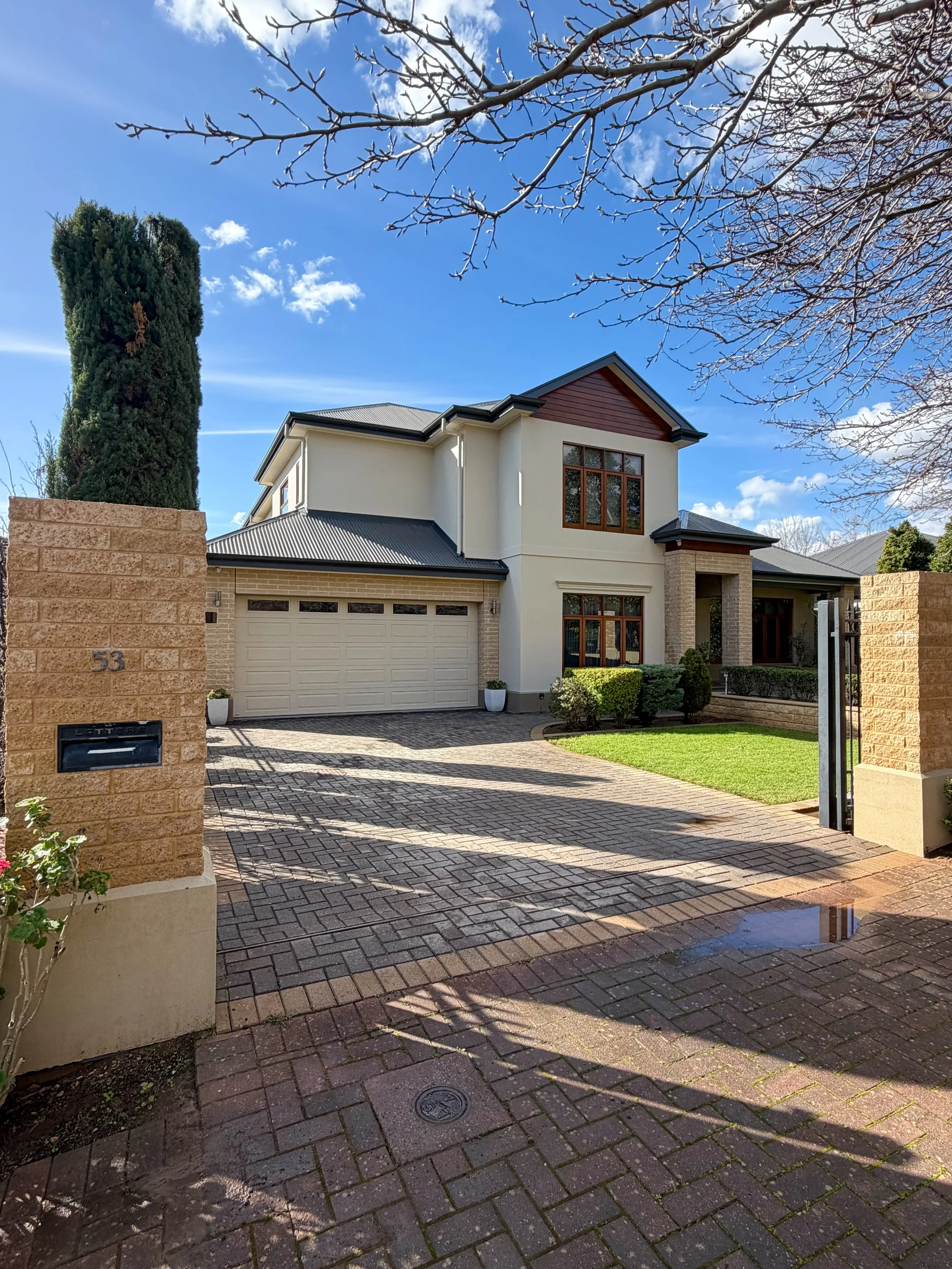 Modern two-story house with a light-colored exterior, large windows, and a gray metal roof, surrounded by a brick-paved driveway and front yard with green grass and bushes. A brick pillar with a mailbox and house number 53 is in the foreground, and a