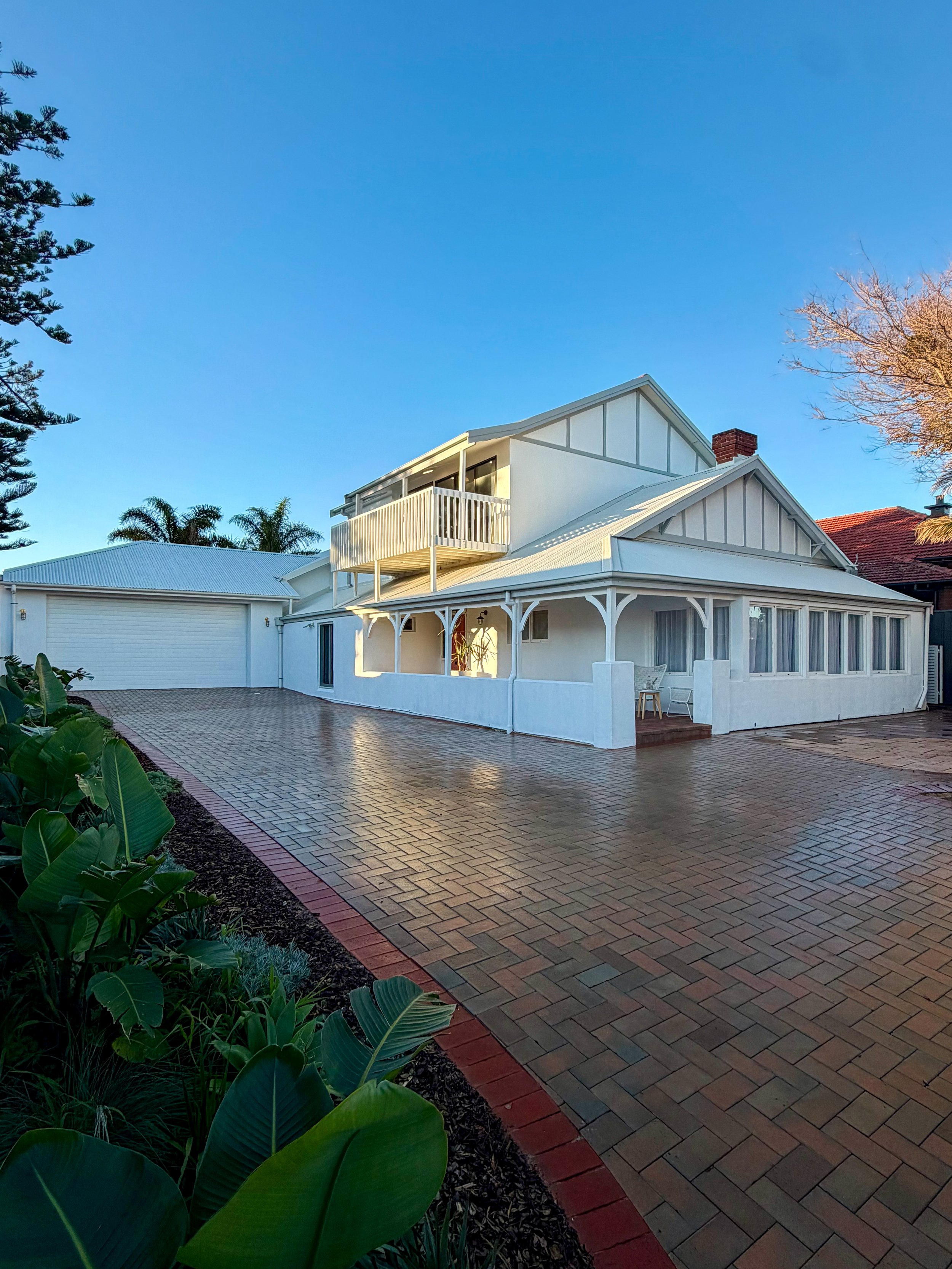 A white house with a porch and balcony, brick driveway, and landscaped garden, under a clear blue sky.