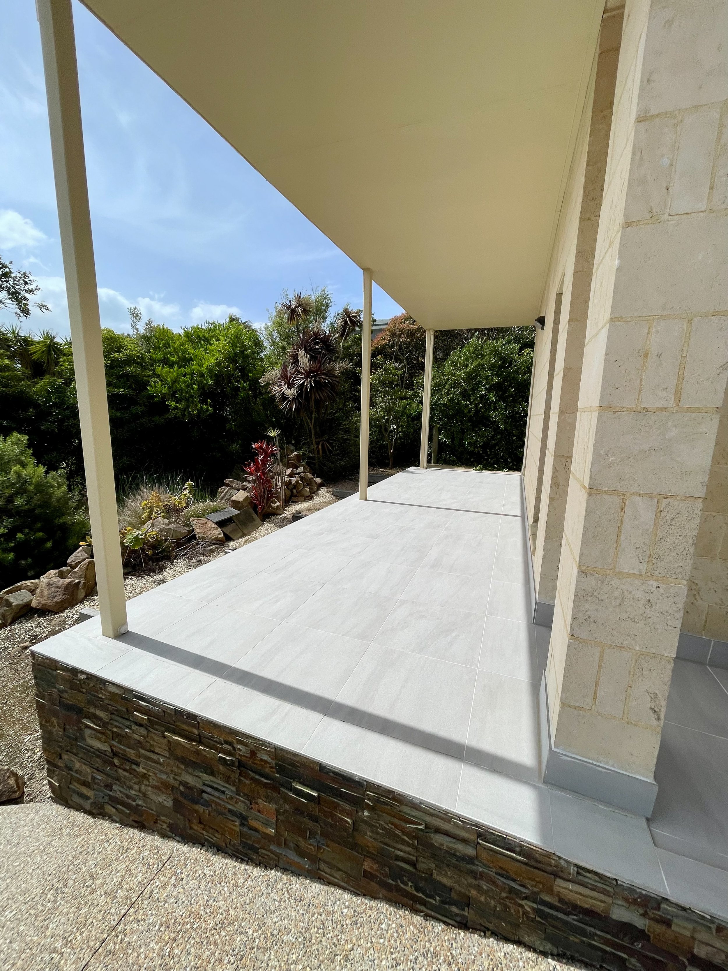 A porch with light gray tiled flooring, stone wall made of multicolored stones, and beige-colored ceiling supported by beige poles. The porch is next to a garden with various green plants and rocks, under a partly cloudy sky.
