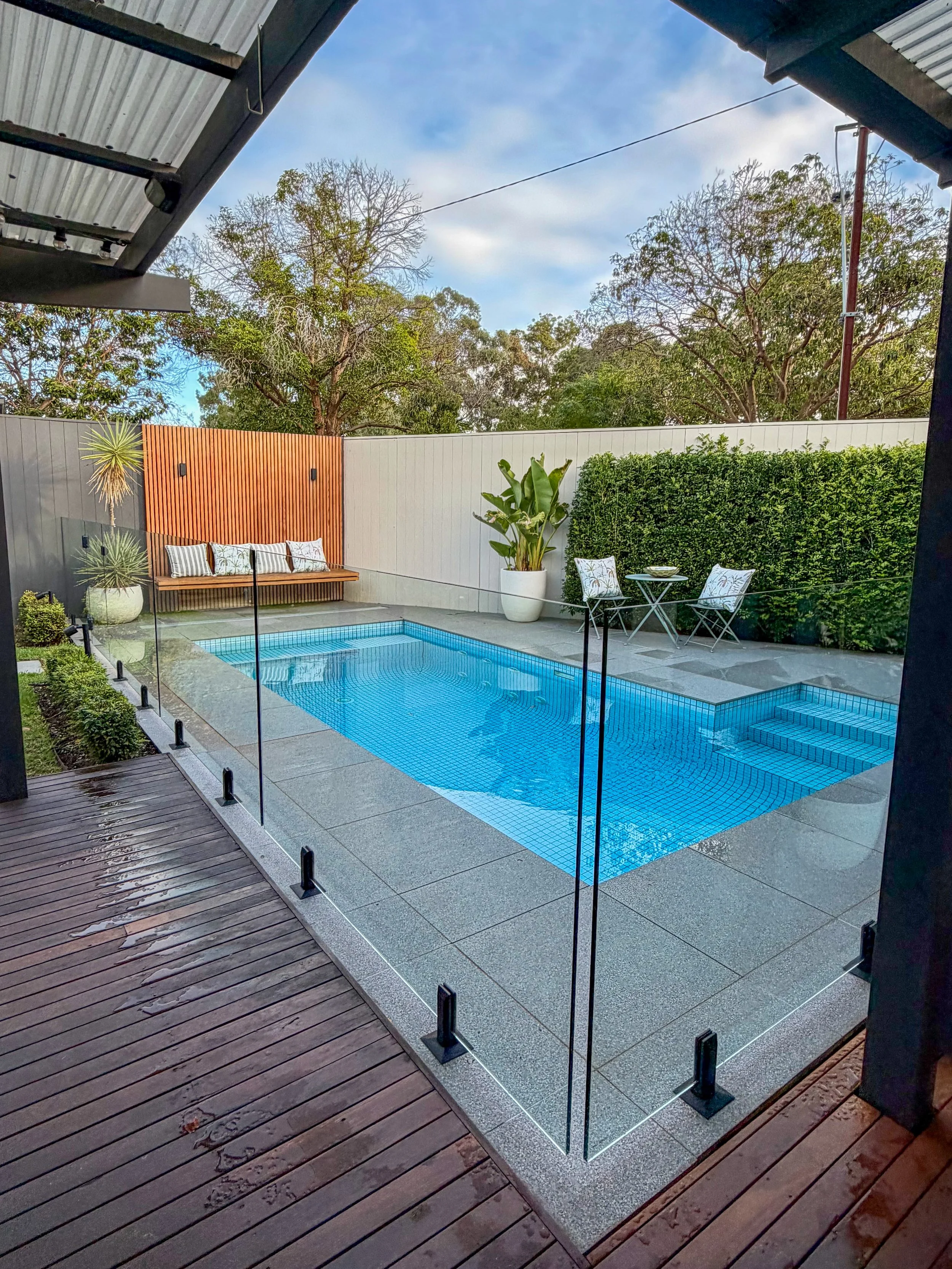 Backyard with small swimming pool, wooden deck, glass fence, outdoor table with two chairs, various potted plants, tall trees, and a white fence under a partly cloudy sky.
