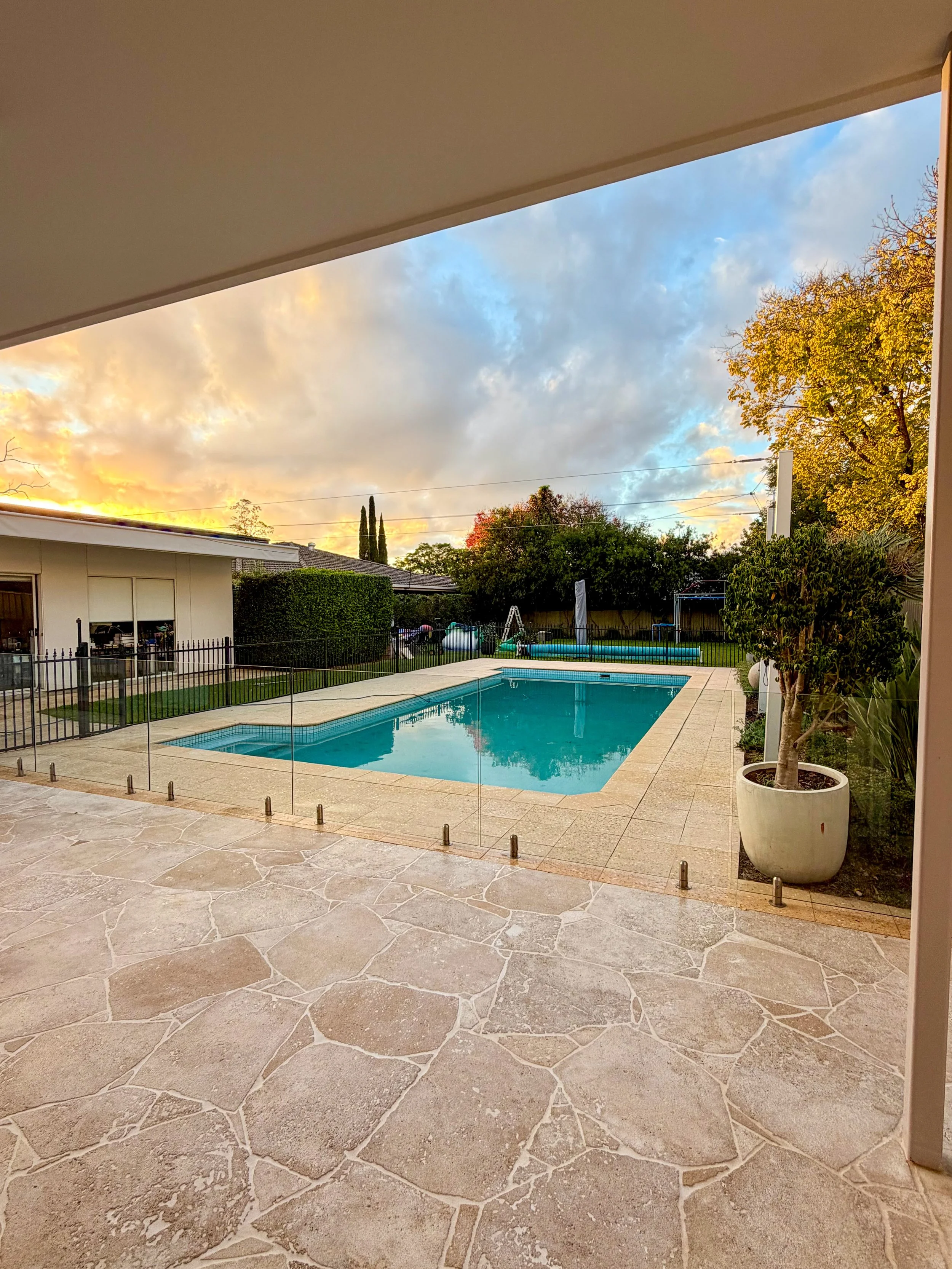View from a patio looking out onto a backyard with a swimming pool, trees, and a sunset sky with clouds.