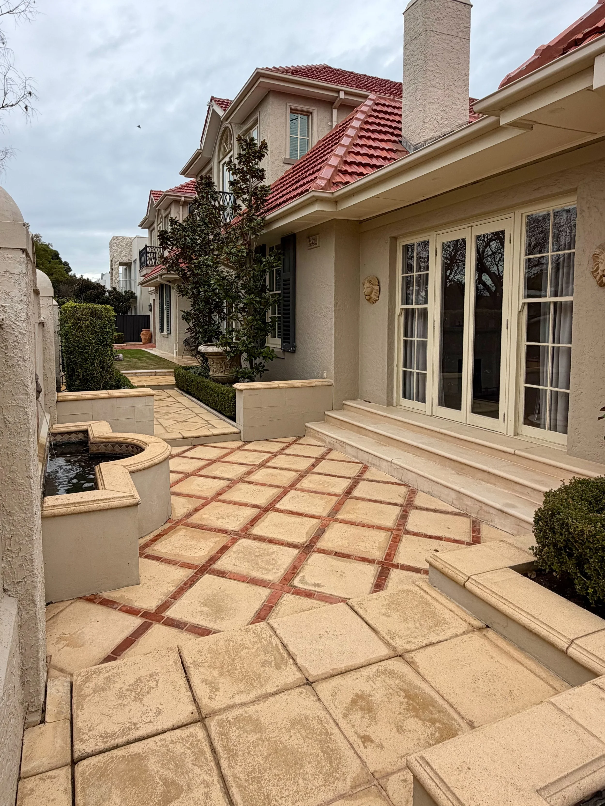 An outdoor patio area of a beige house with a red-tiled roof, large windows, and a small waterfall feature surrounded by pavers and greenery.