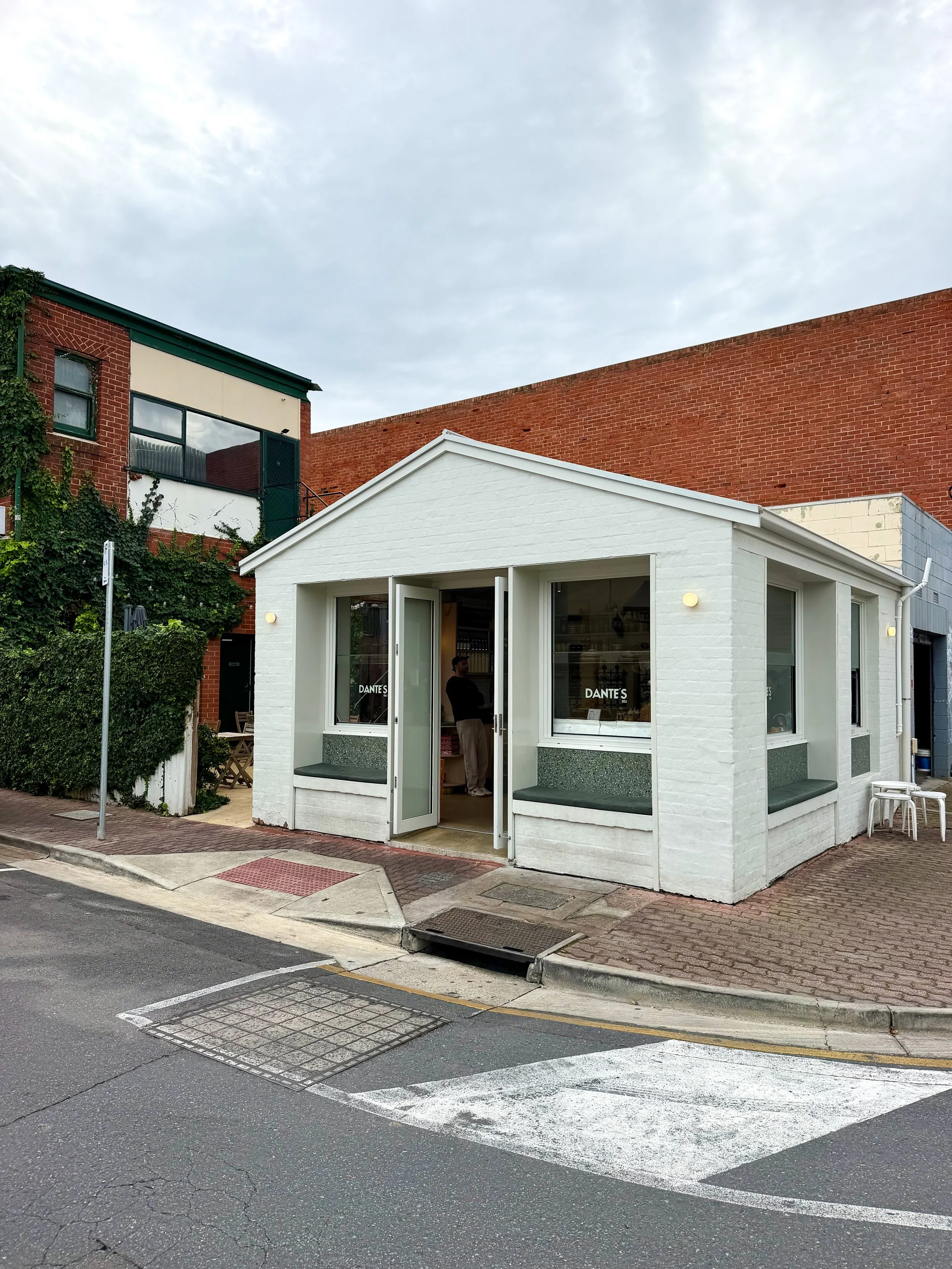 Street corner with a small white building housing a cafe called Dante's, a man inside the cafe, a brick building behind it, street signs, and marked parking spaces on the road.