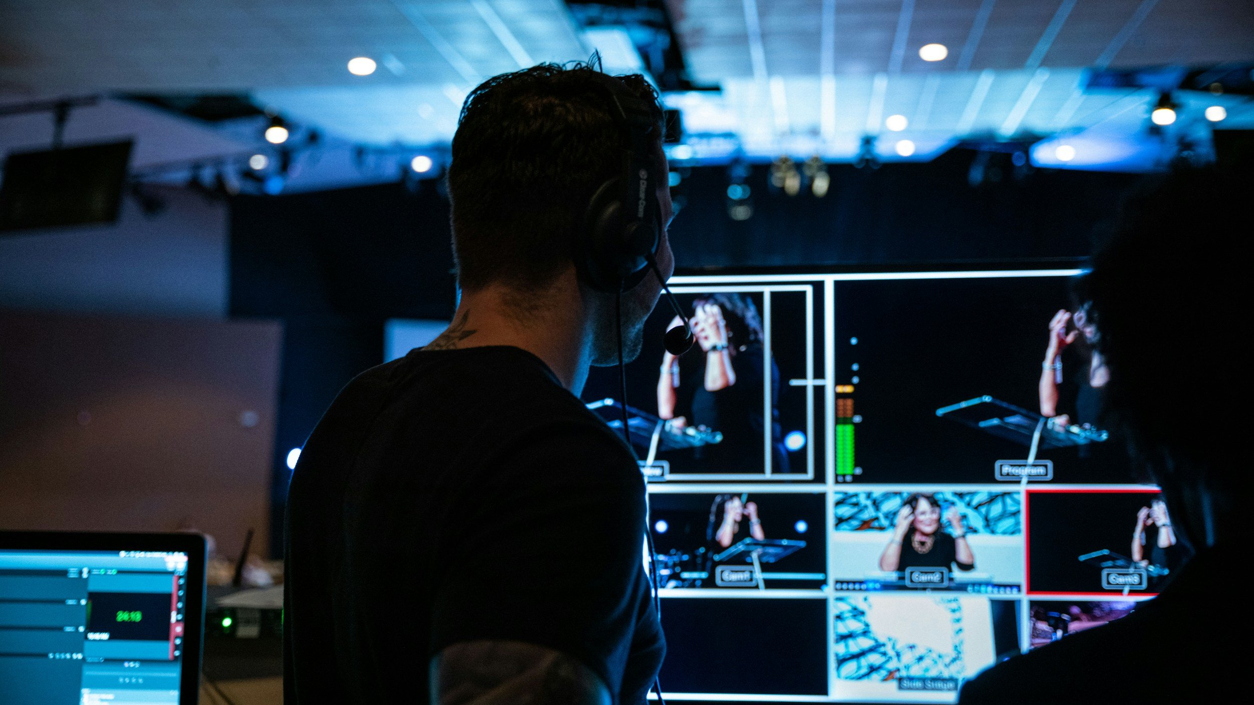 A cameraman operating a professional video camera at an indoor event with blue lighting and a seated audience in the background.