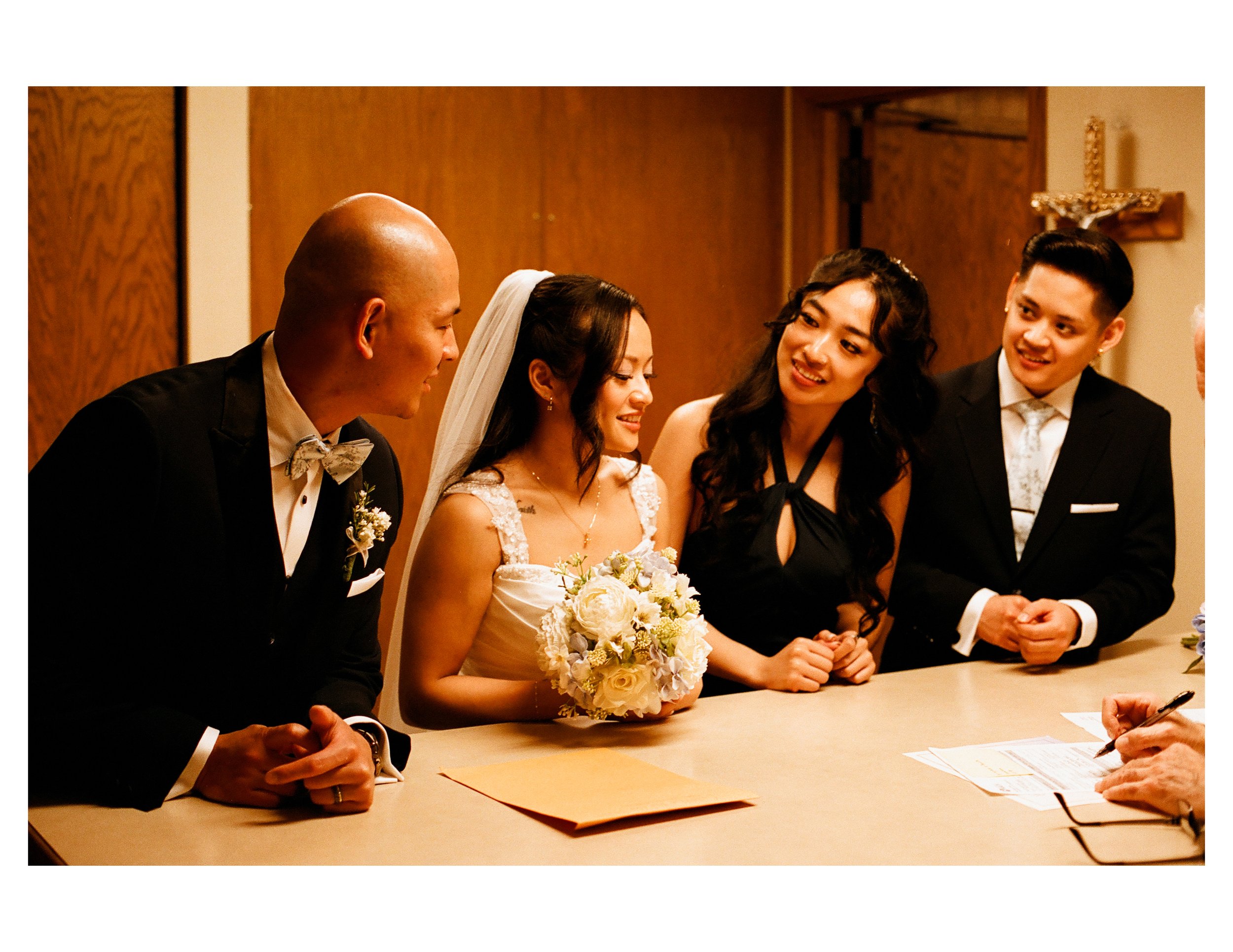 A wedding ceremony with the bride and groom signing documents, surrounded by friends and family in a wooden-paneled room with a cross on the wall.