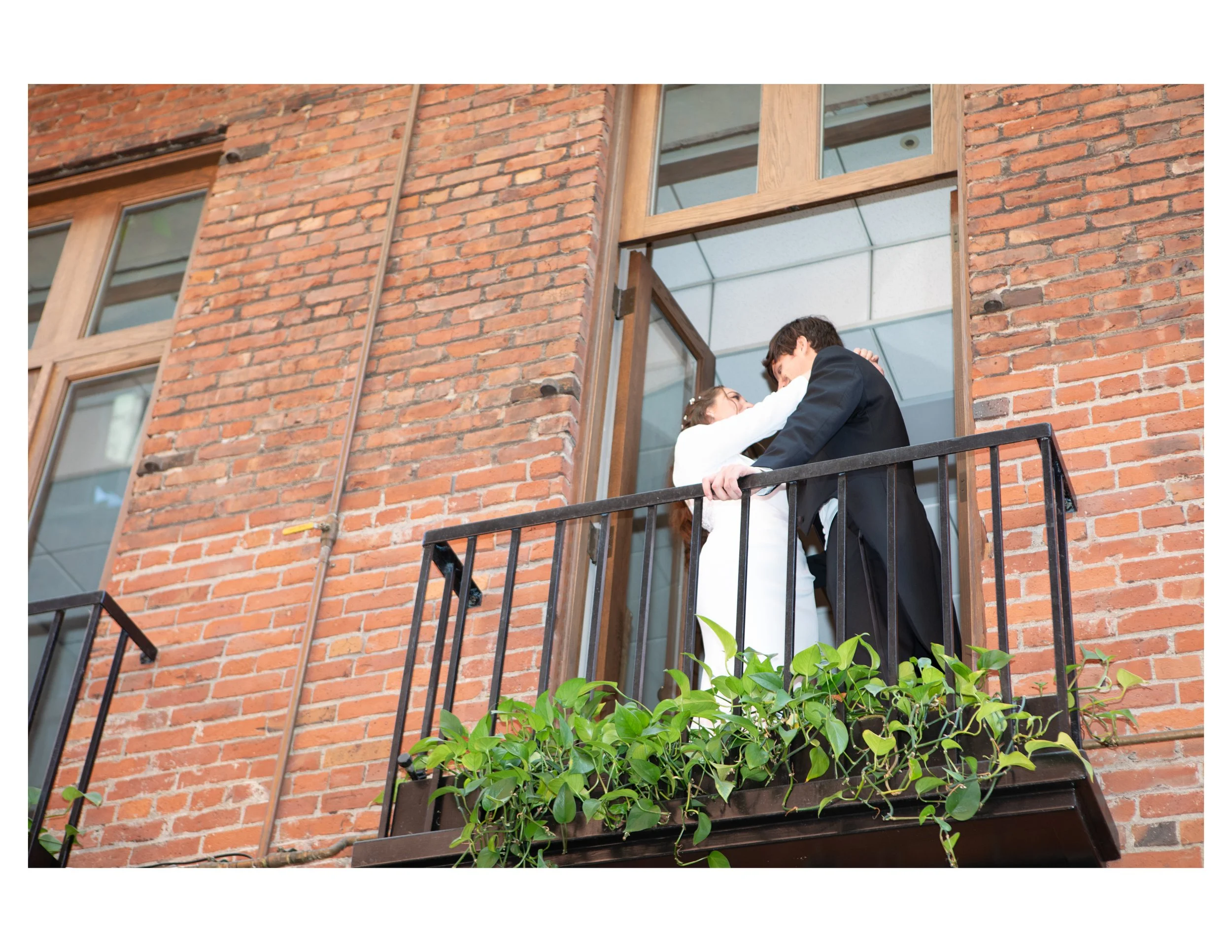 A young couple with the man in a suit and the woman in a white dress sharing a romantic moment on a balcony of a brick building.