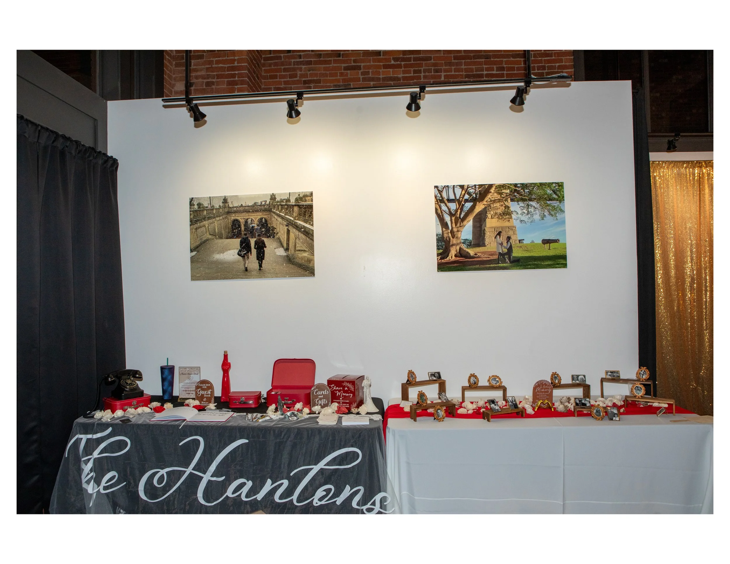 Indoor photography exhibition with photographs and memorabilia display, including a table with framed pictures, signs, and decorative items, set against white walls with track lighting, black curtains on the left, brick wall at the top, and a golden 