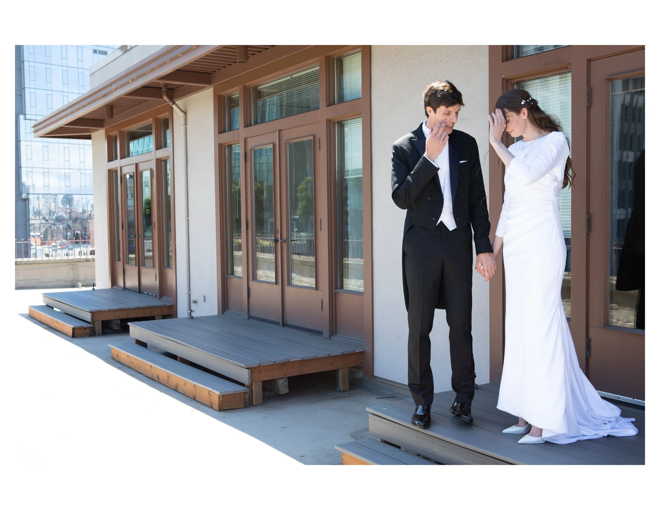 A bride and groom holding hands and touching foreheads on an outdoor wooden terrace with modern buildings in the background.