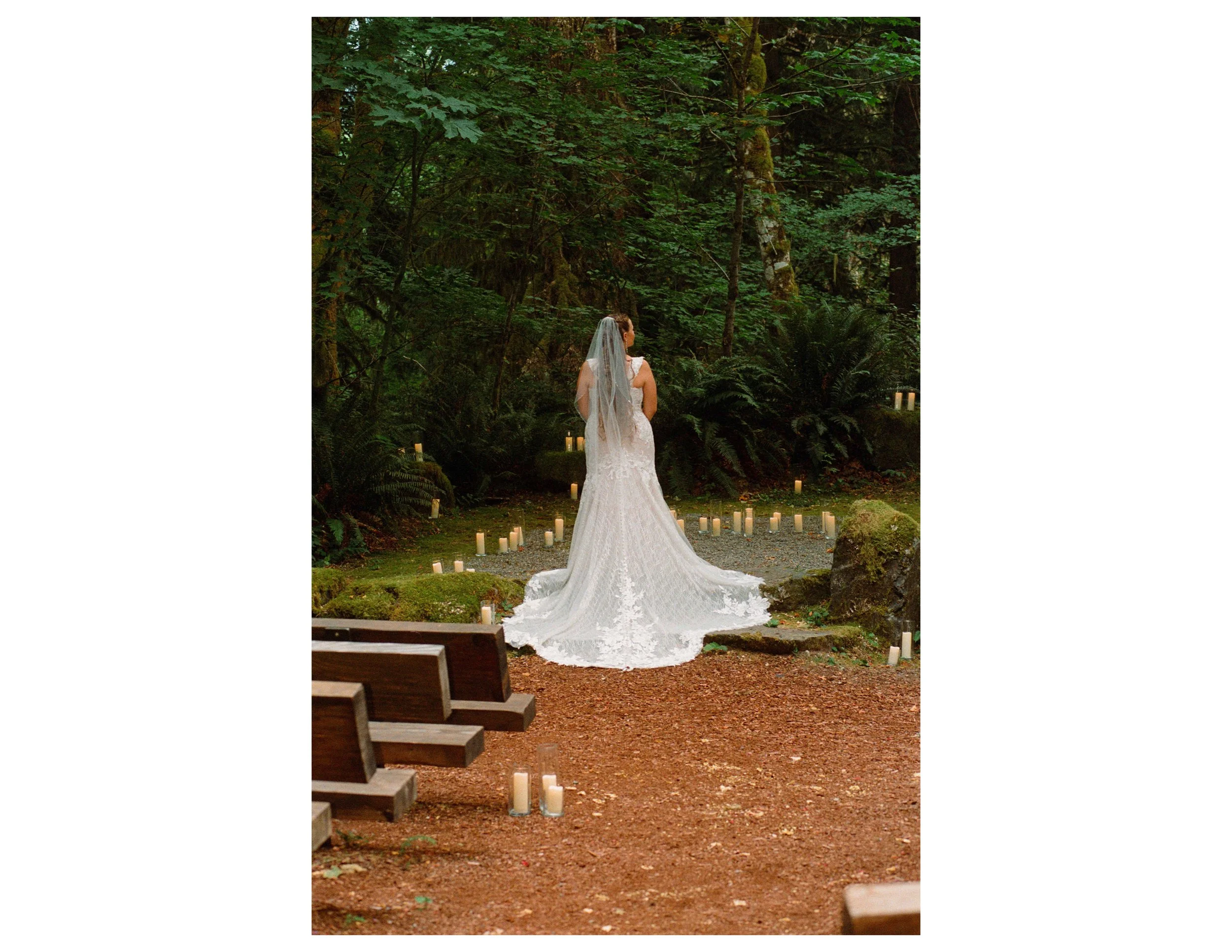 A bride in a white wedding dress and veil standing outdoors in a forested area, surrounded by candles on the ground.