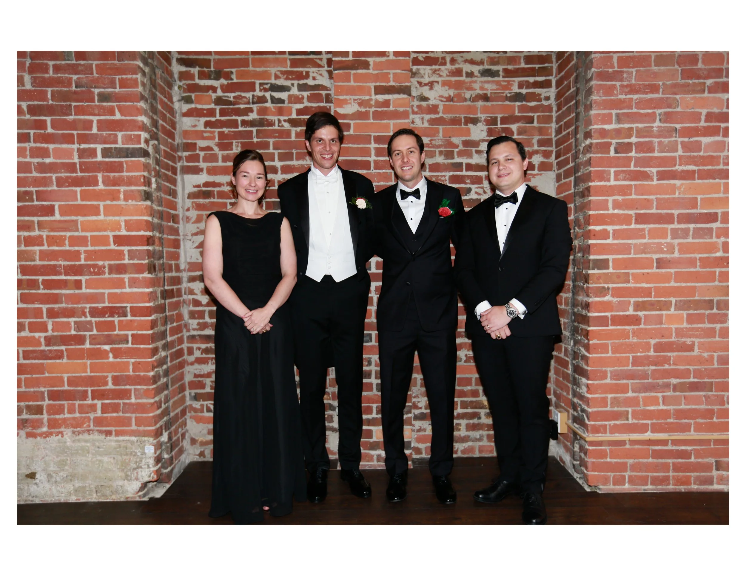 Four people in formal attire standing against a brick wall, smiling at the camera.