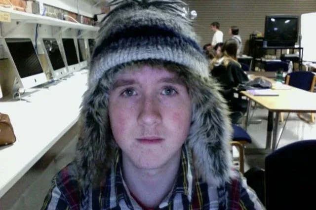 Young man with a fur-lined winter hat in a computer lab or classroom setting.