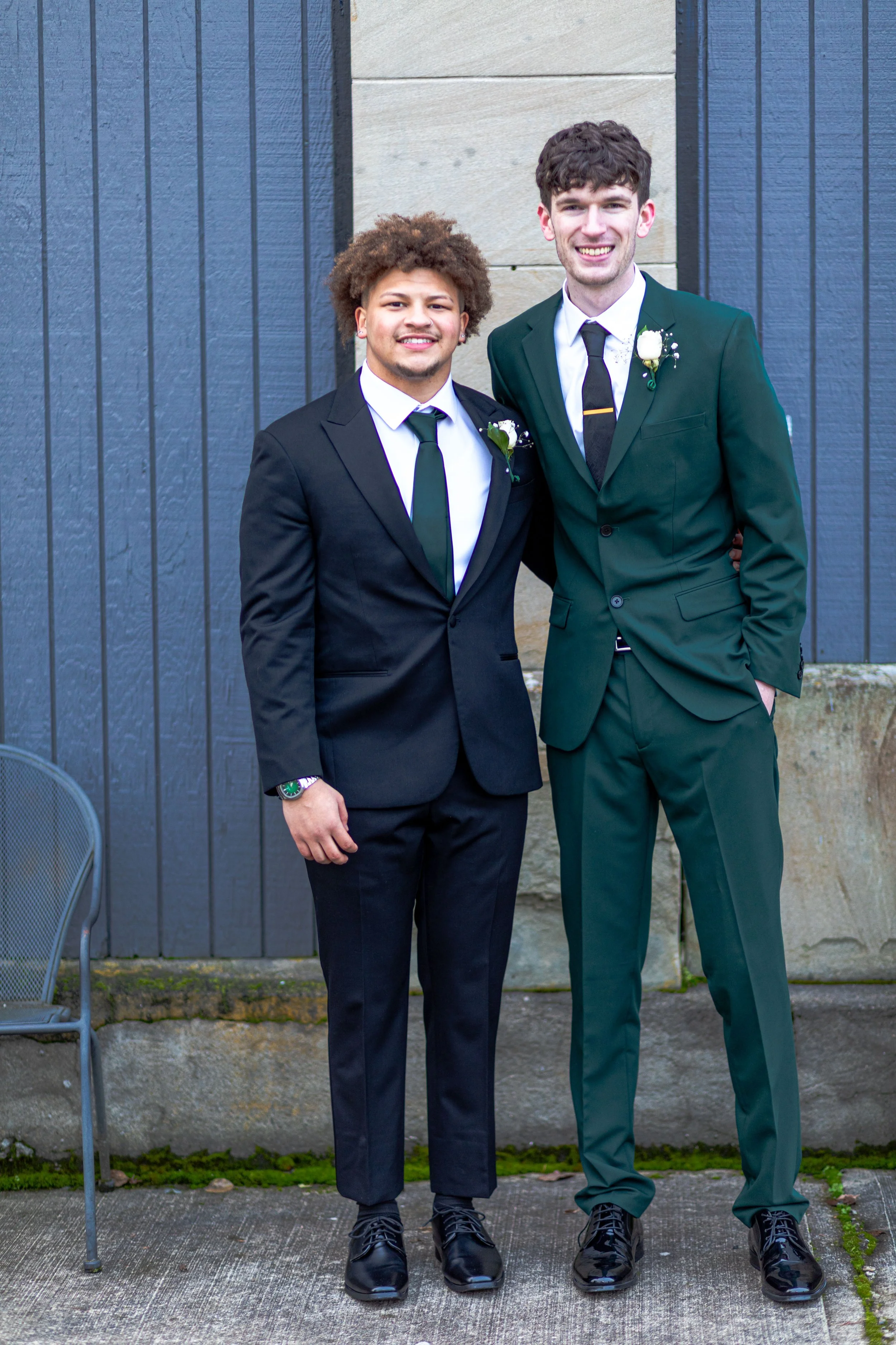 Two men in suits standing outside, smiling, with blue and beige wall background.