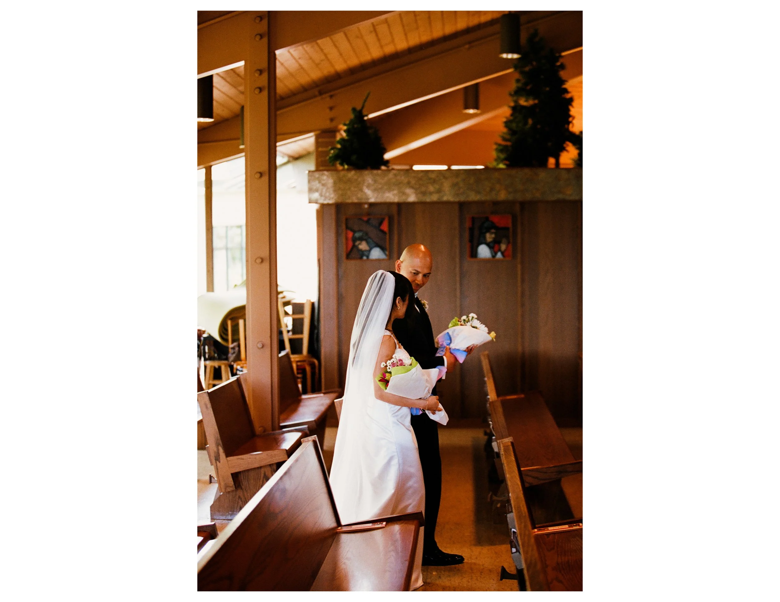 A couple, possibly newlyweds, standing inside a church or similar venue, sharing a moment with the bride in a white dress and veil holding a bouquet, and the groom in a dark suit, with religious artwork on the wooden wall behind them.