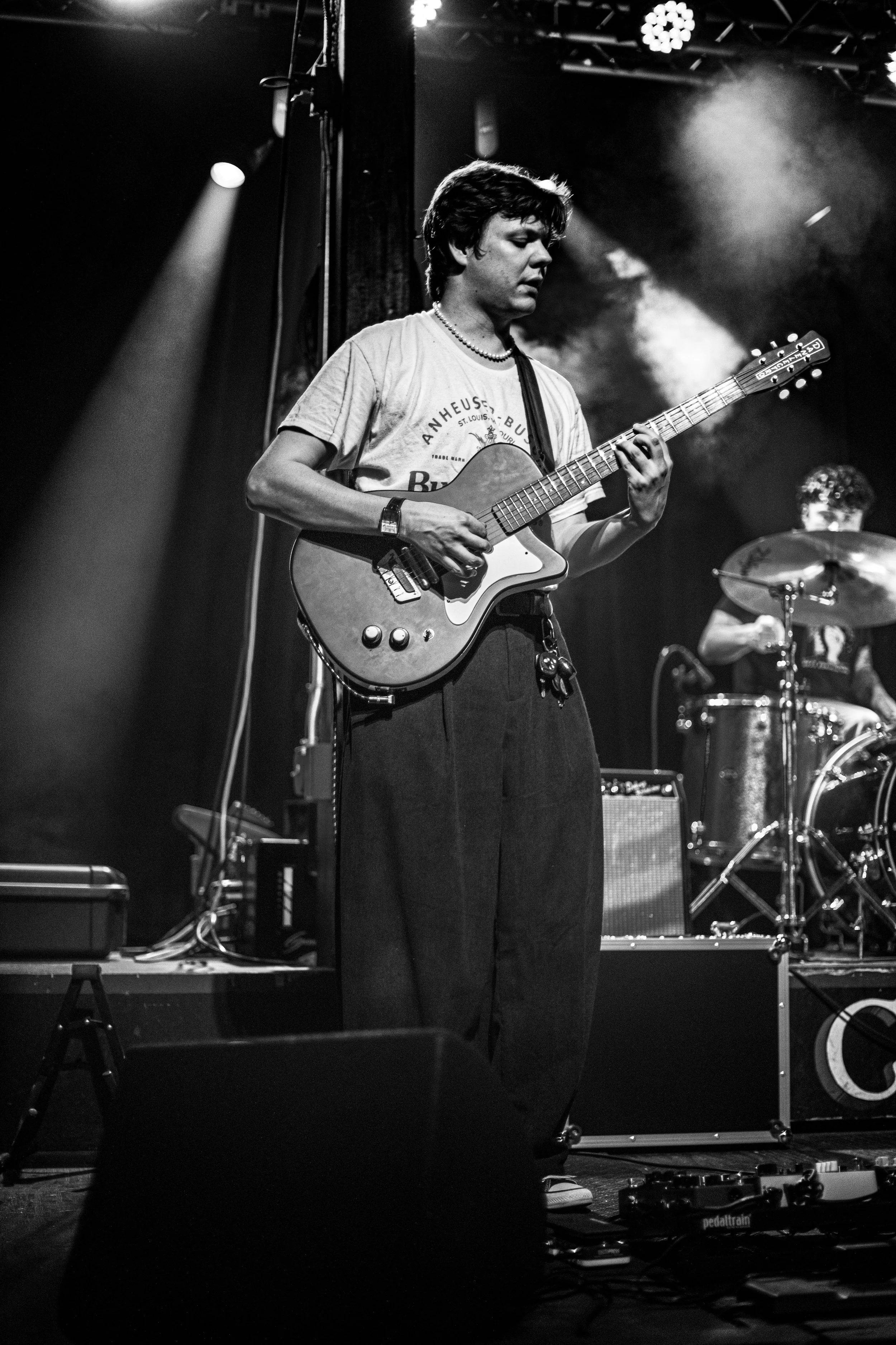 A young man playing an electric guitar on stage during a live music performance, with a drummer in the background and stage lighting overhead.