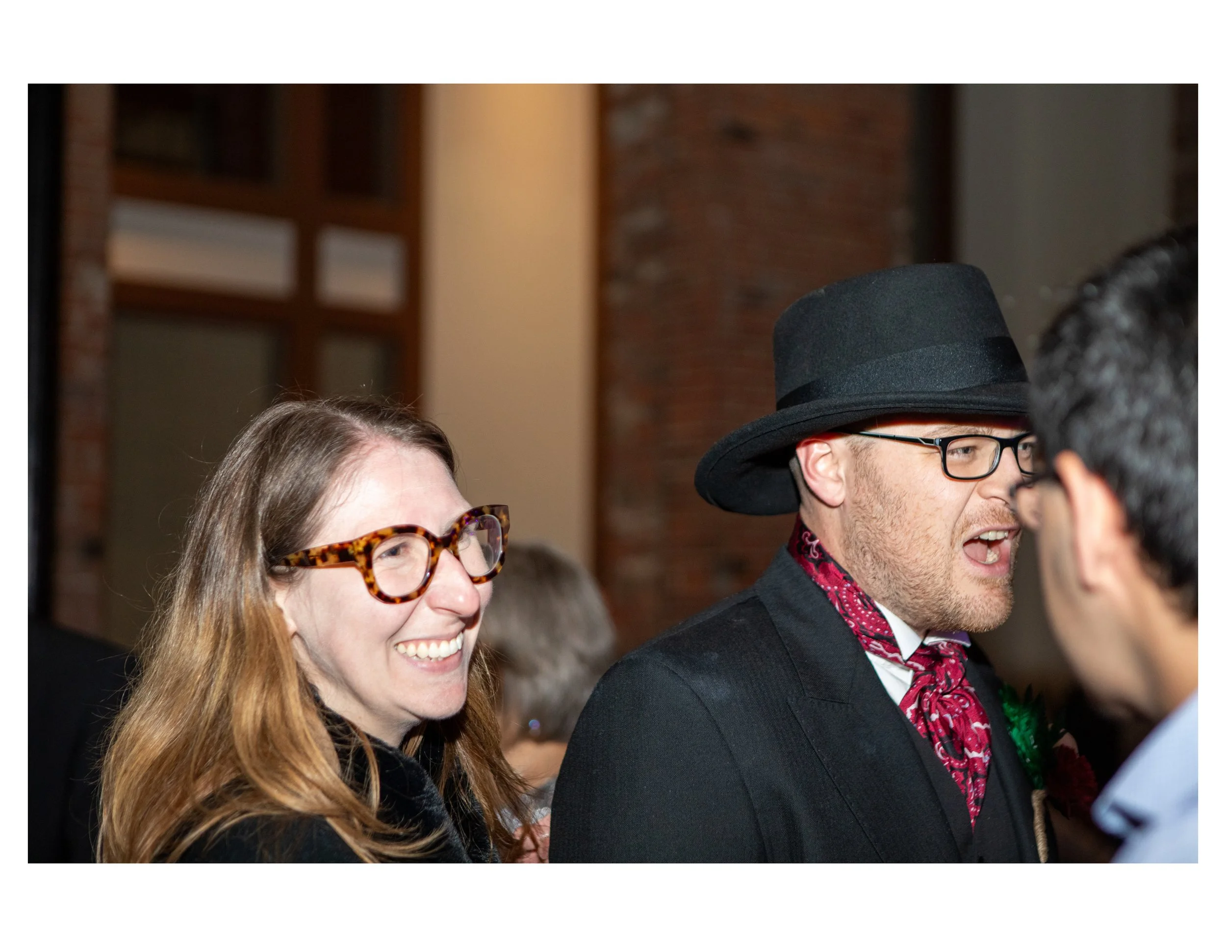 Two people smiling at an event, the woman wearing tortoiseshell glasses and the man wearing a black hat and glasses.