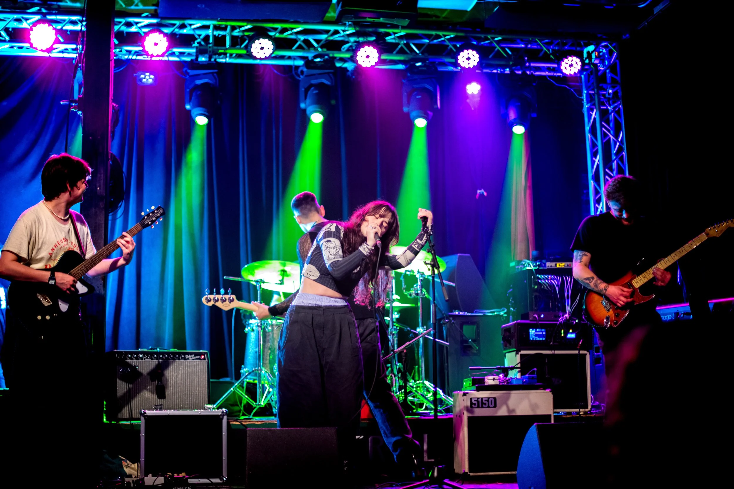 A band performing live on stage with colorful lighting, including female singer with microphone, guitarist on left, bassist on right, and drummer in the background.