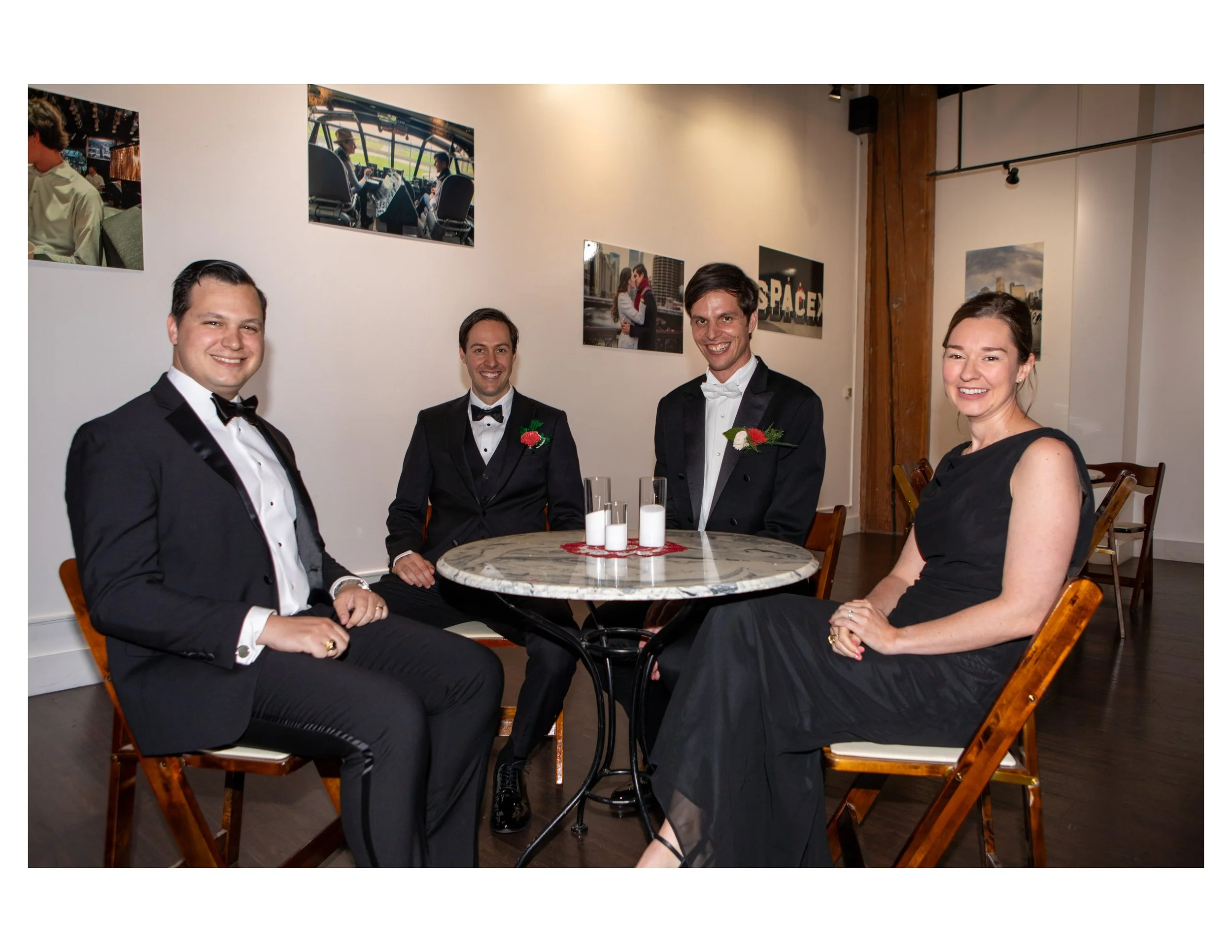 Four people dressed in formal tuxedos and evening dresses sitting and standing around a table with candles in a decorated room, with photos on the wall in the background.