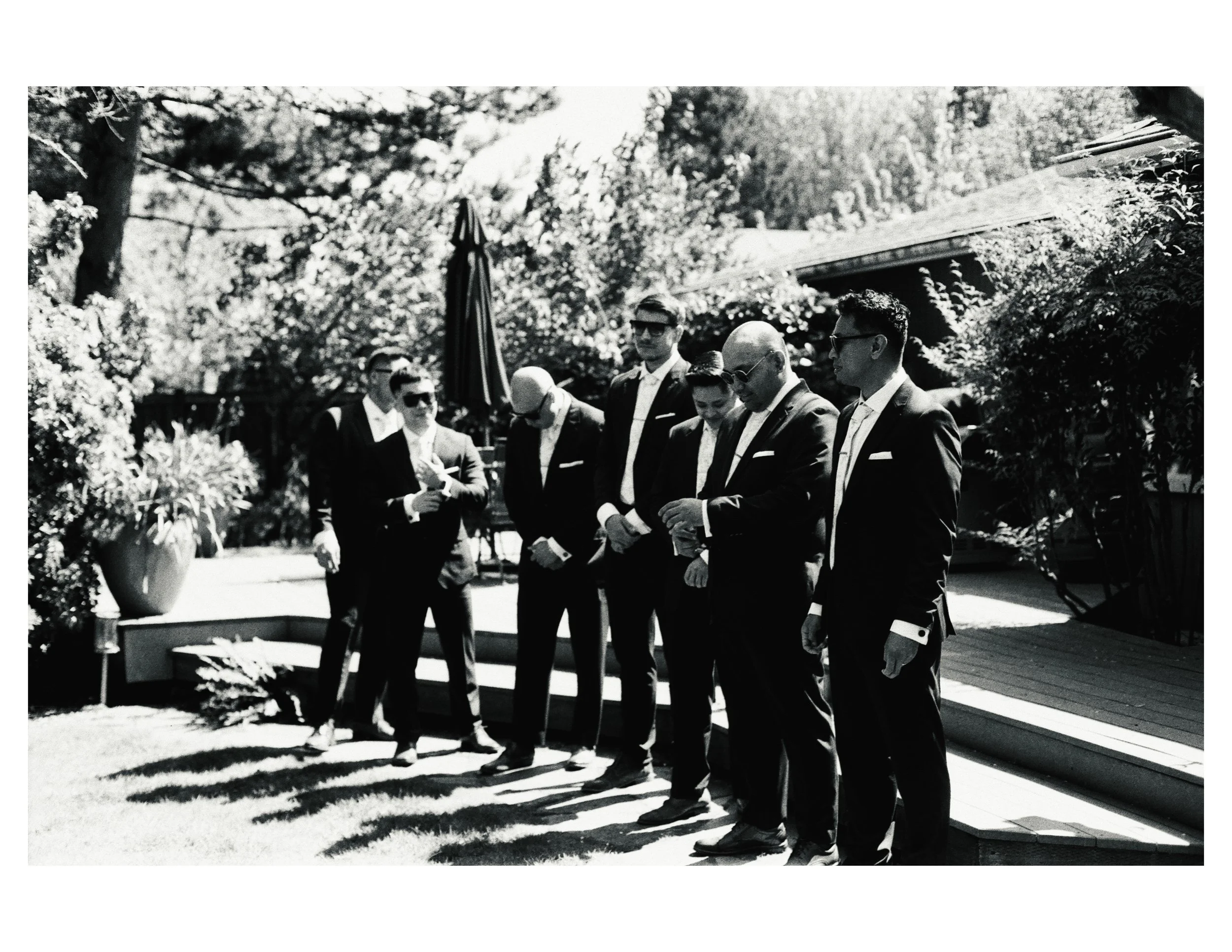 A group of people dressed in formal suits standing outdoors in a garden or park setting, with trees and a house in the background, possibly at a wedding or formal gathering.