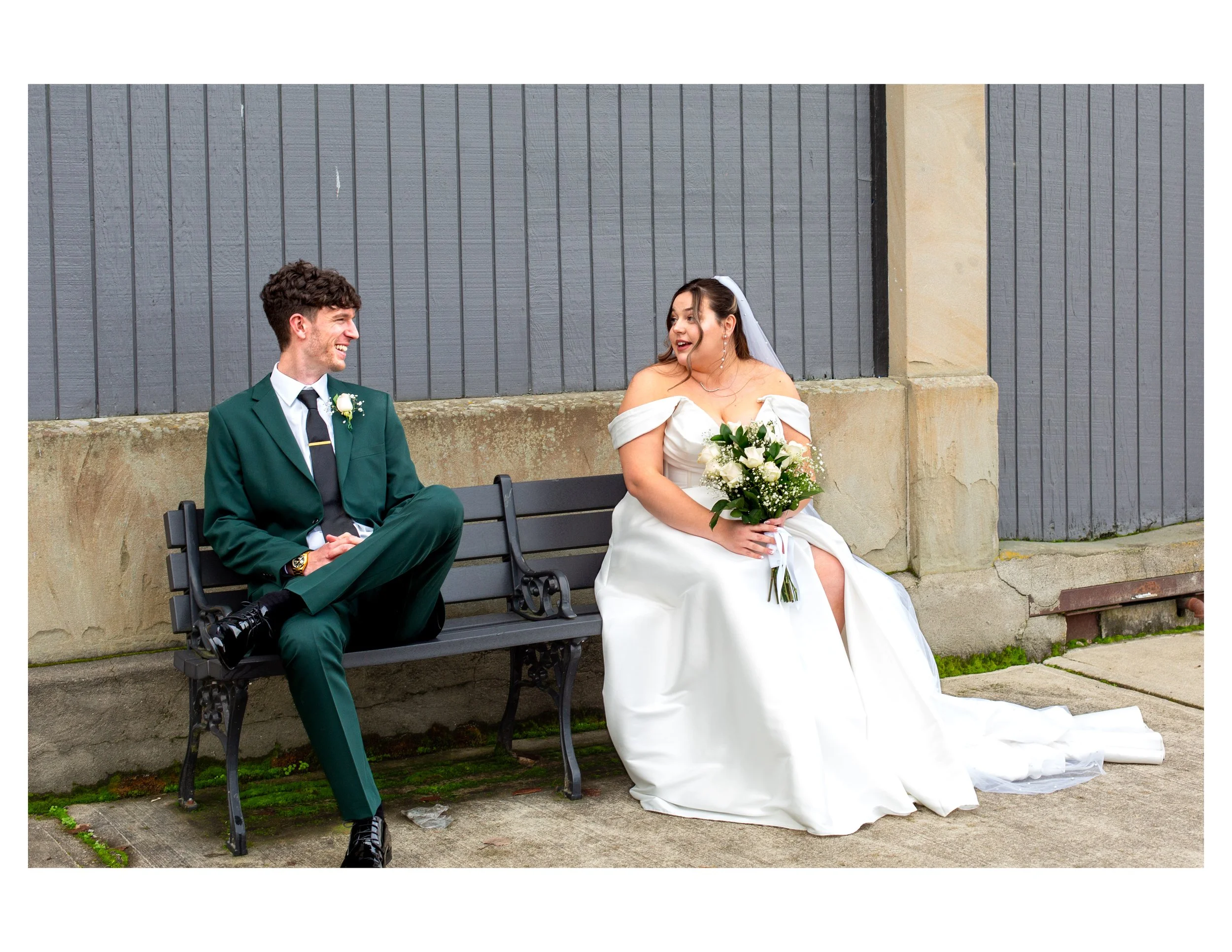 A bride and groom sitting on a bench outdoors, smiling and engaging in conversation, with a gray wooden fence and stone column in the background.