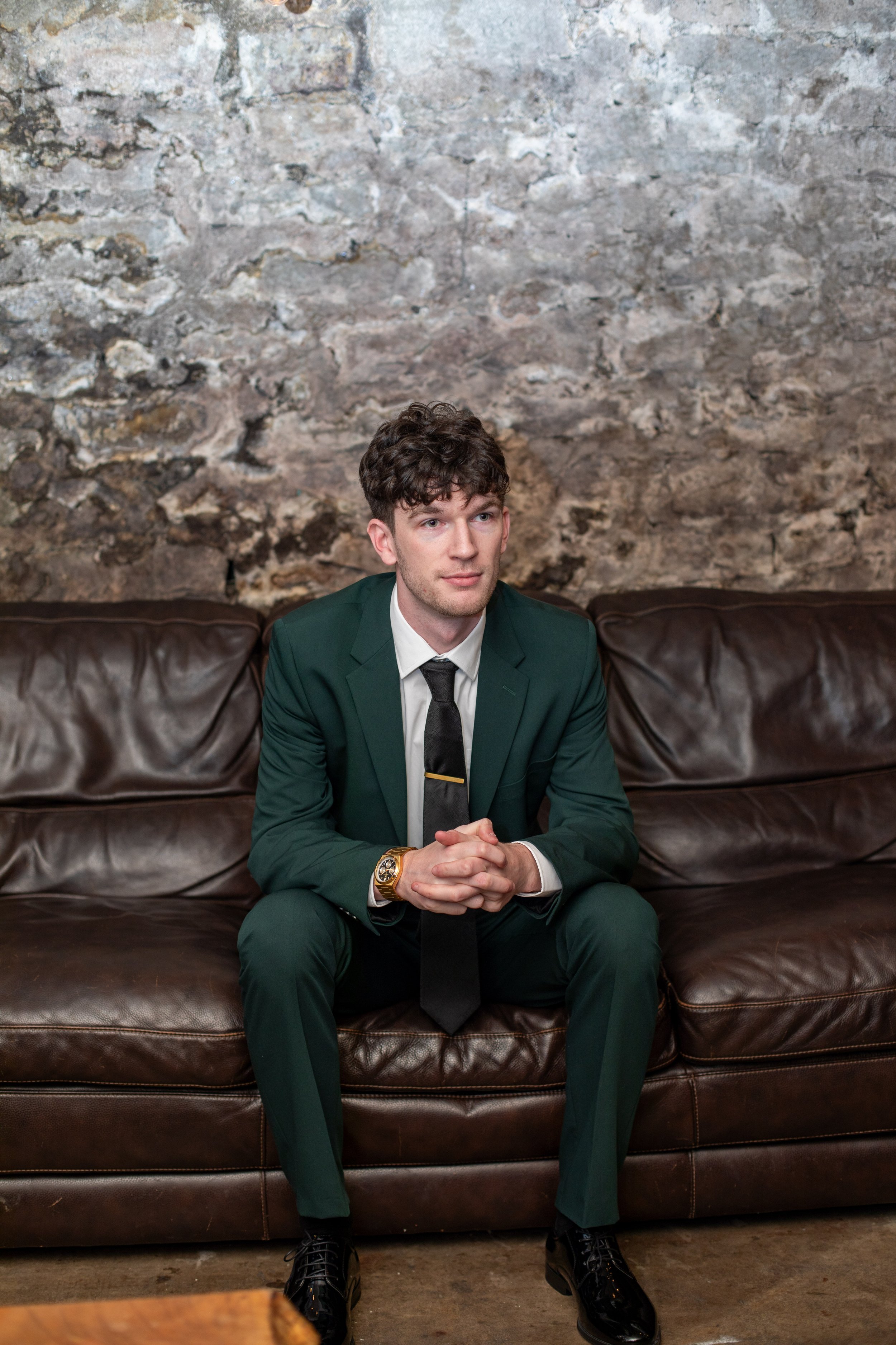 A young man in a dark green suit, white shirt, and black tie sitting on a brown leather sofa in front of a textured stone wall.