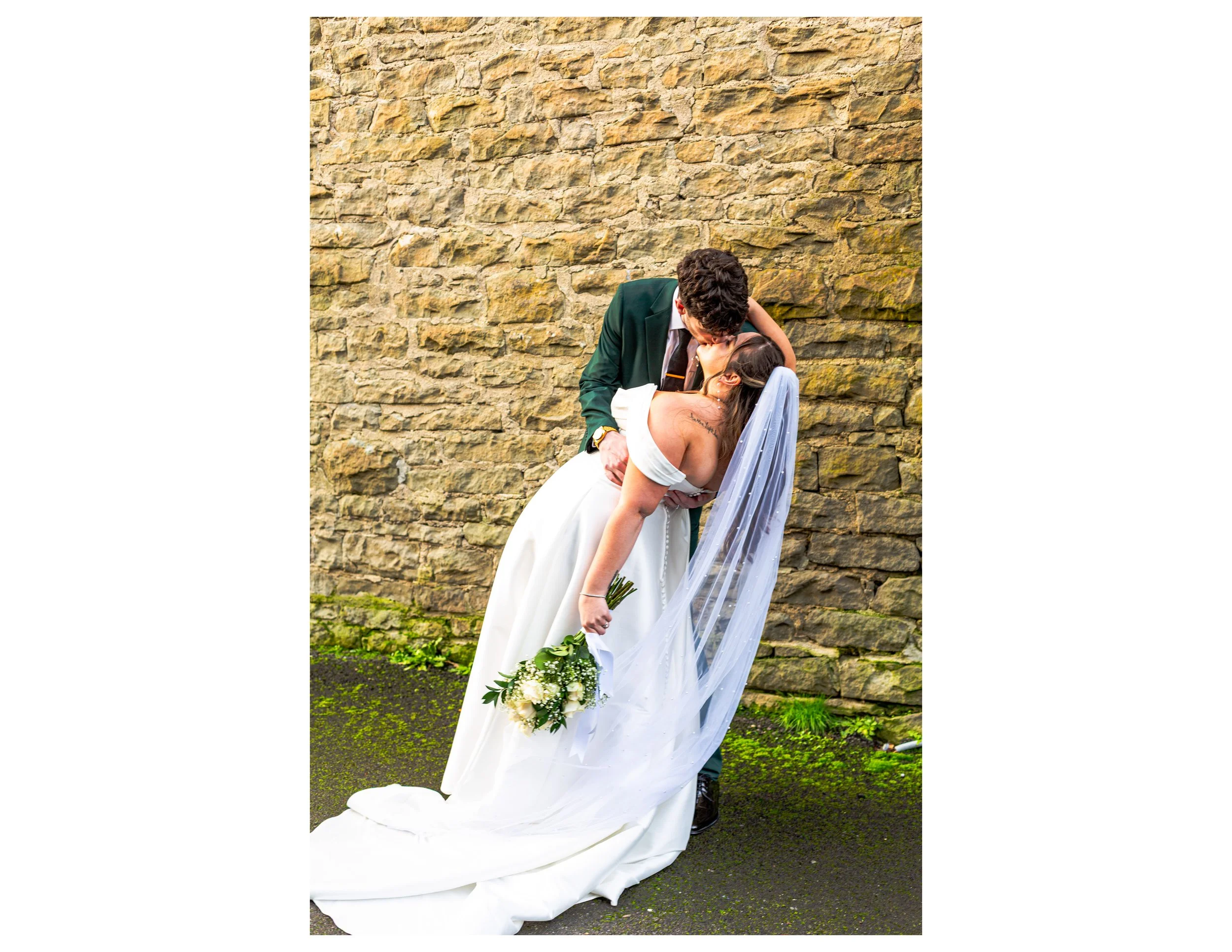A newlywed couple sharing a kiss outdoors against a stone wall. The bride is in a white wedding gown holding a bouquet of white and green flowers, and the groom is in a dark suit. The bride is leaning backward as the groom dips her for a kiss.