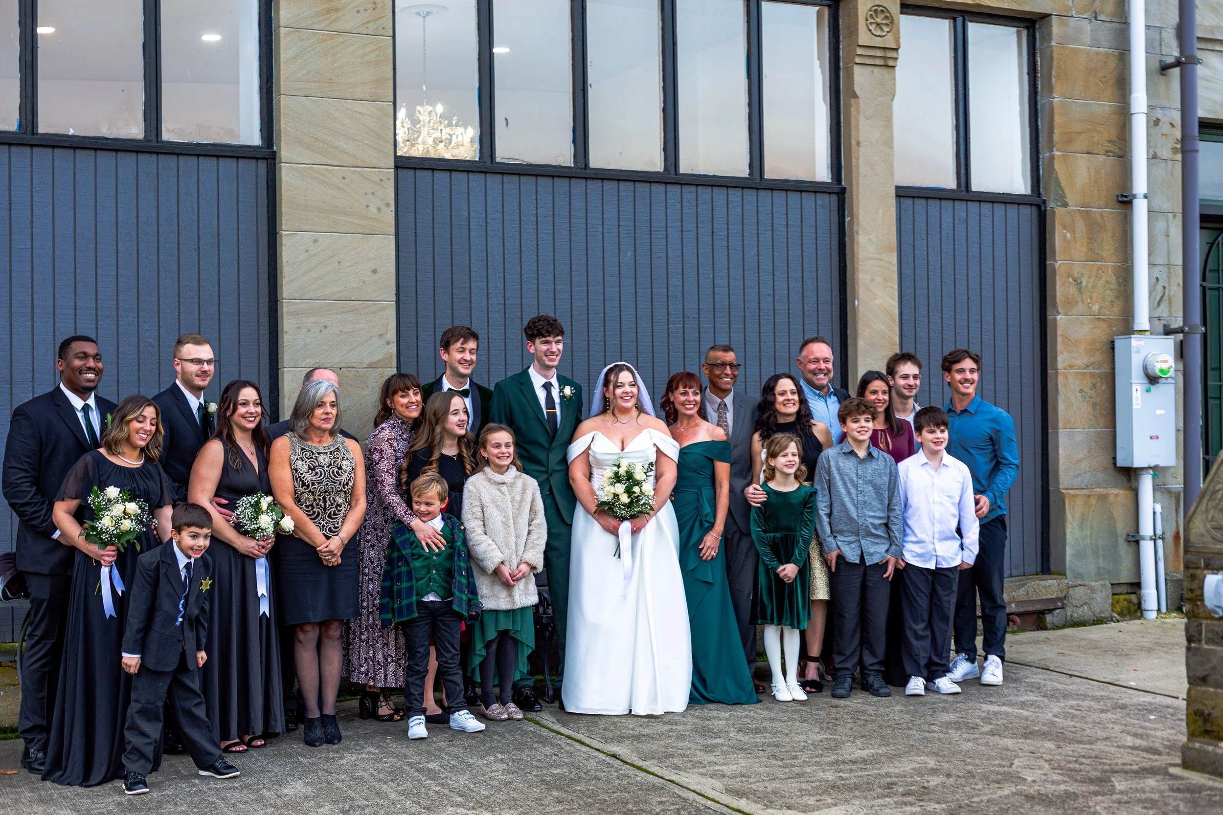A group of people in formal attire, including a bride and groom, standing outdoors in front of a building with large windows, celebrating at a wedding.