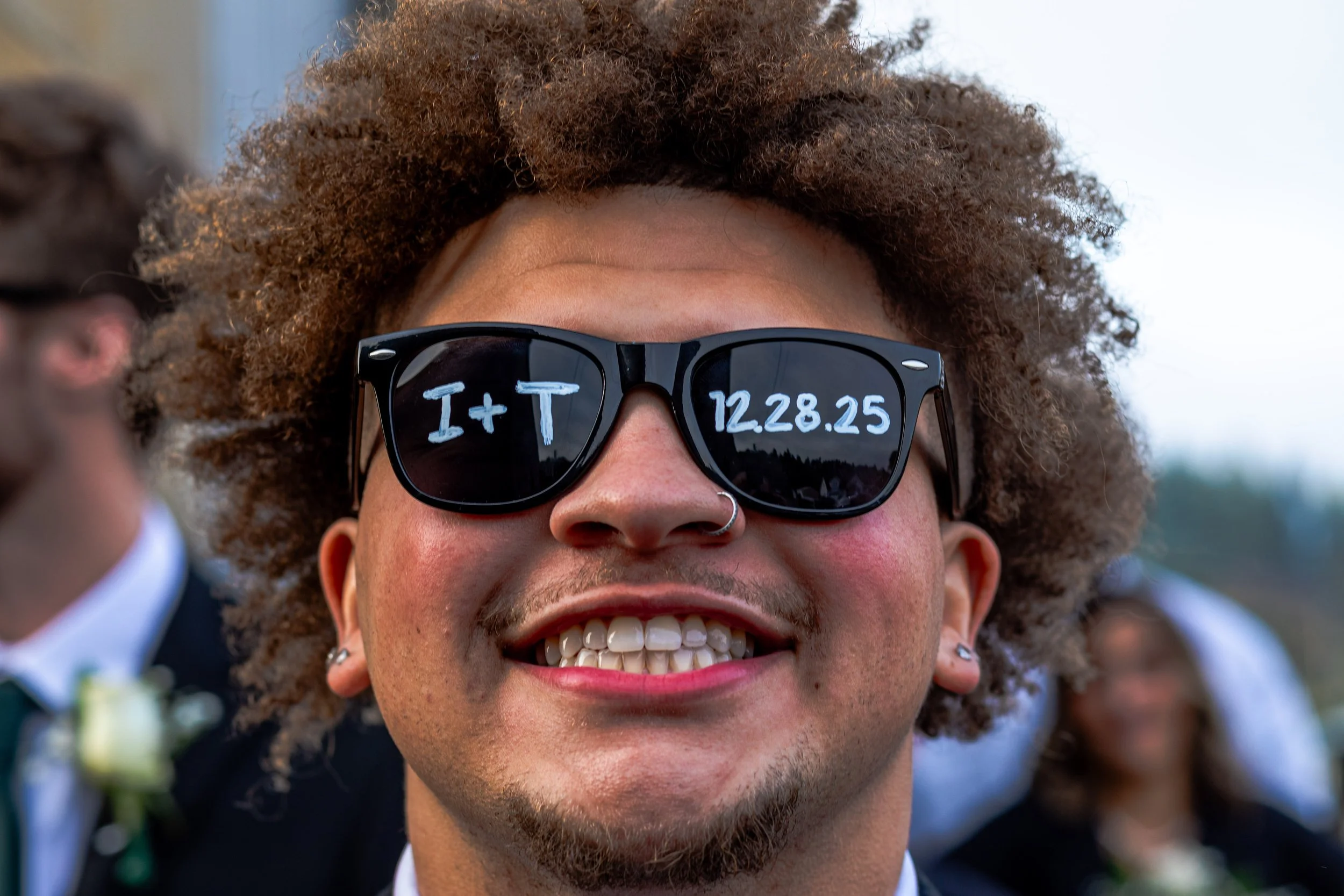 A person with curly hair wearing black sunglasses with writing on the lenses that say 'I + T' and the date '12.28.25', smiling outdoors at a wedding or event.