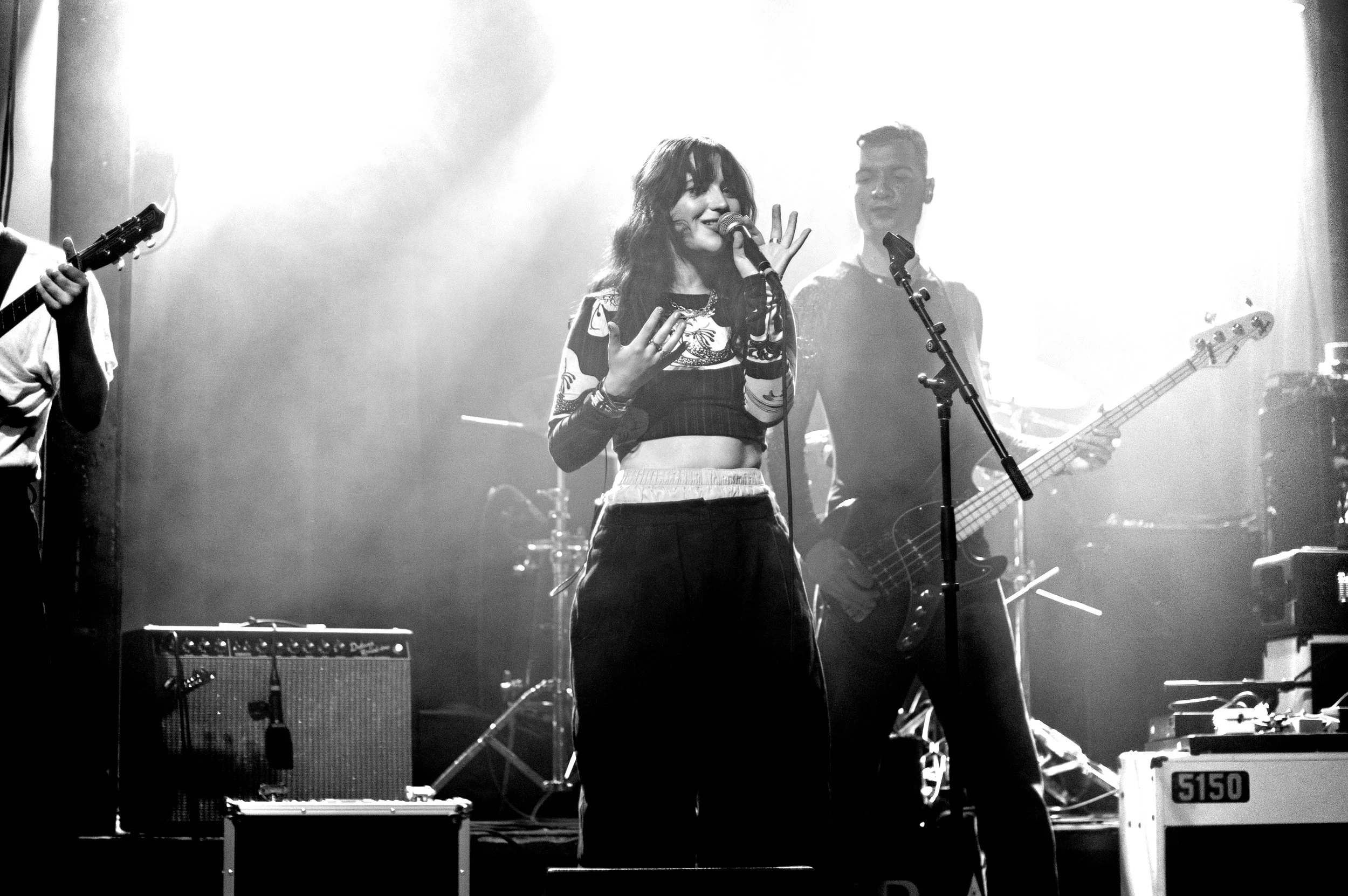 A woman singing into a microphone on stage with a guitarist and drum set in the background.