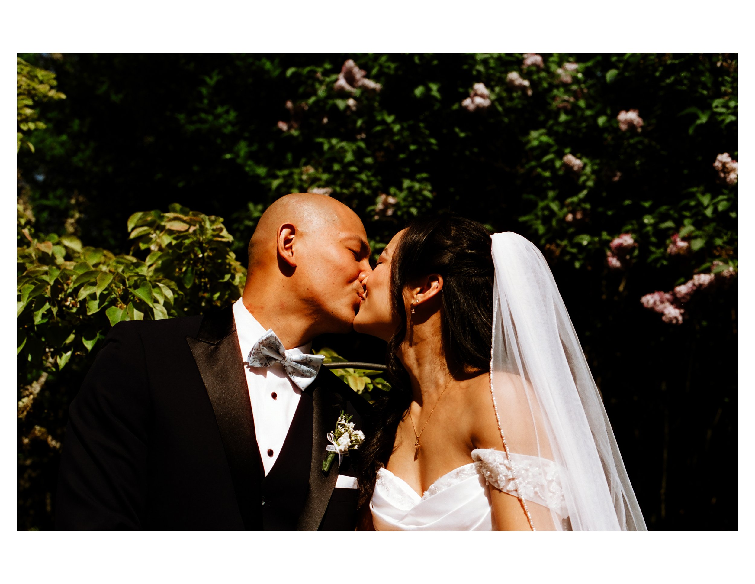 A bride and groom share a kiss outdoors, with greenery and pink flowers in the background, during a wedding ceremony.