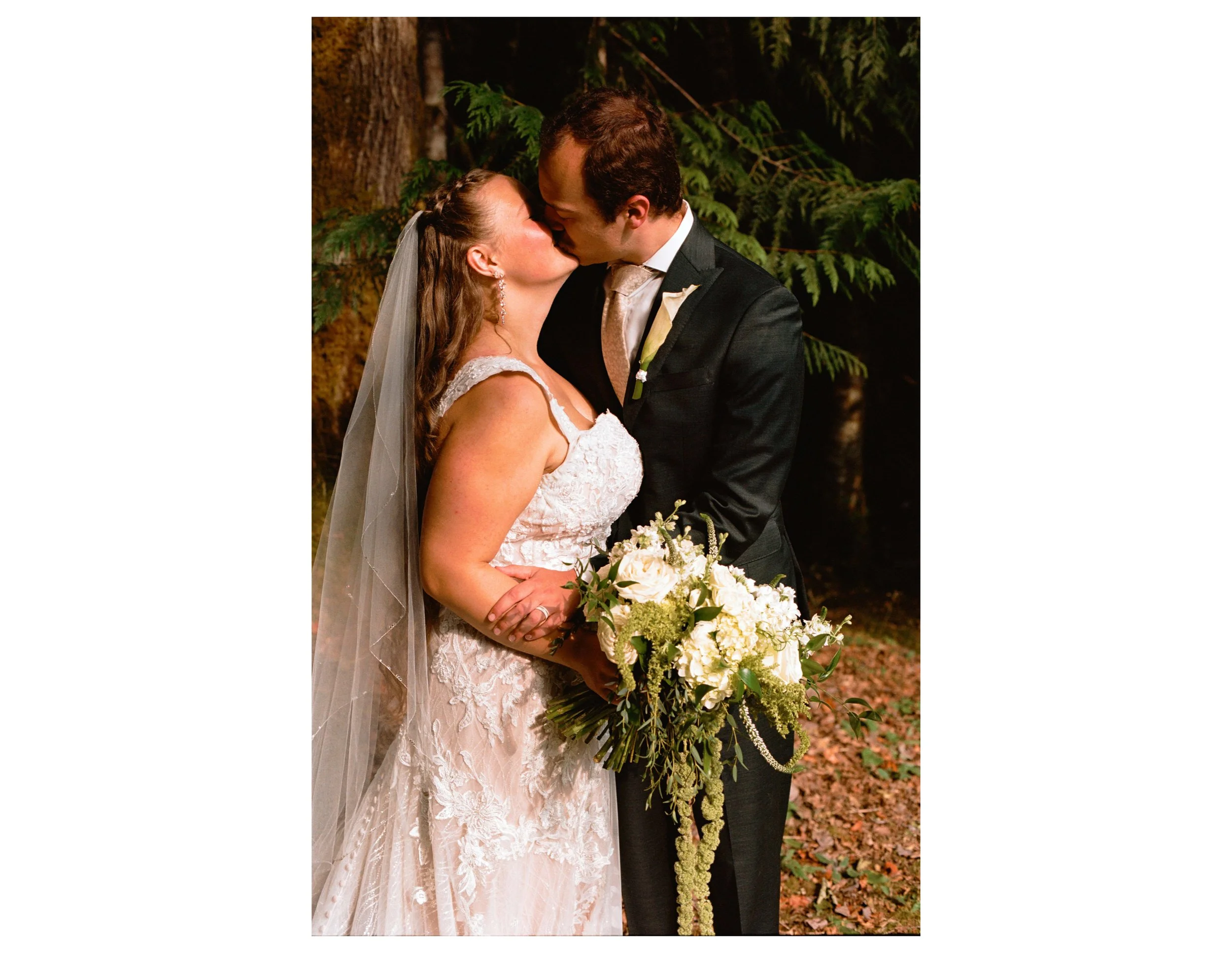 A bride and groom sharing a kiss outdoors at night, with green foliage and tree trunks in the background. The bride wears a white lace wedding dress with a veil, and the groom is dressed in a dark suit with a white shirt and light-colored tie. They h