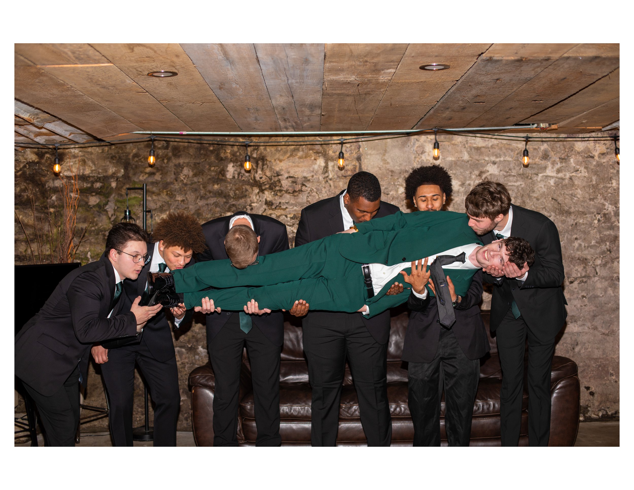 Group of men in suits participating in a horse kissing event in a rustic indoor setting with a stone wall and hanging Edison bulbs.
