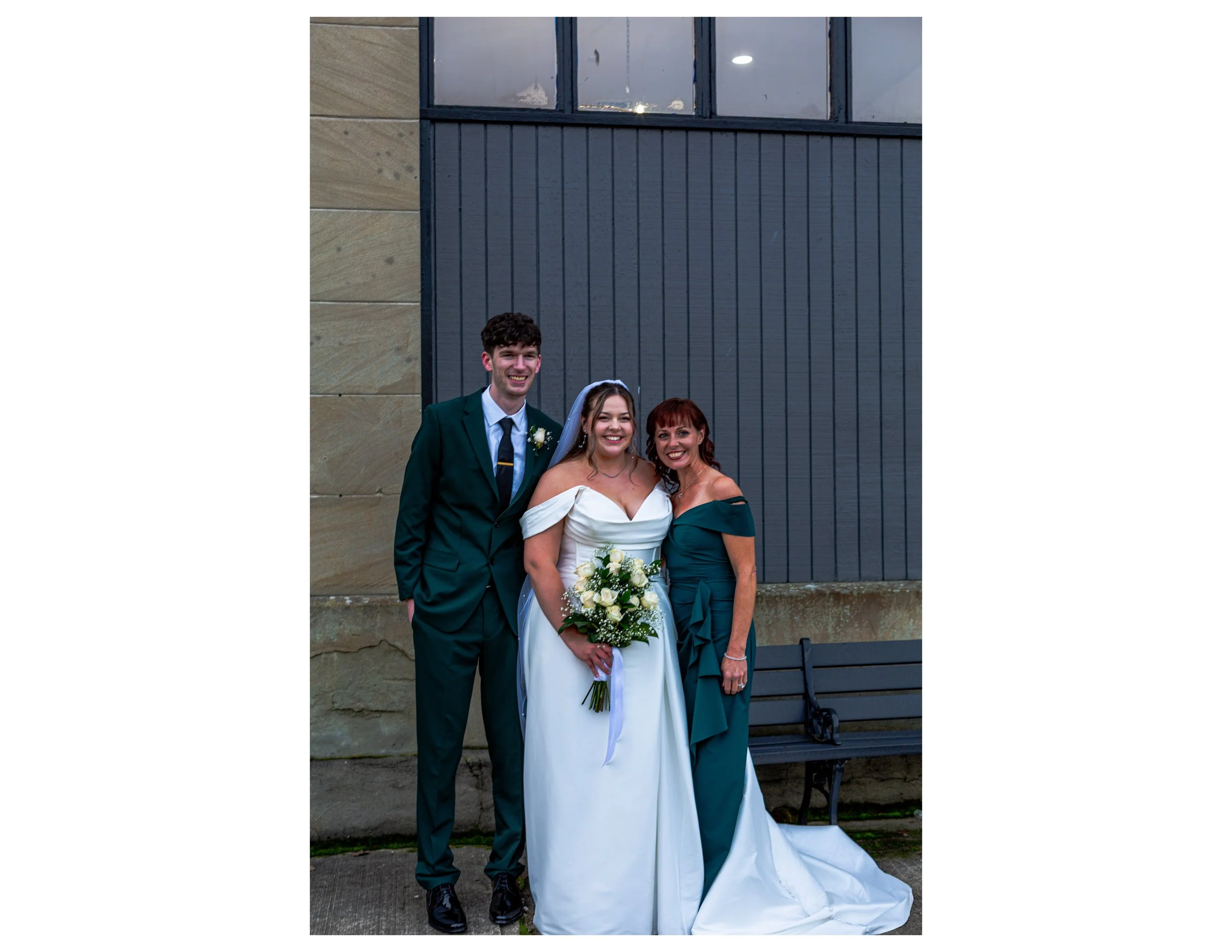 A wedding portrait of three smiling people, including a man in a suit with a boutonniere, a woman in a white wedding dress holding a bouquet, and a woman in a dark green dress, standing outdoors in front of a gray wooden wall and beige stone wall.
