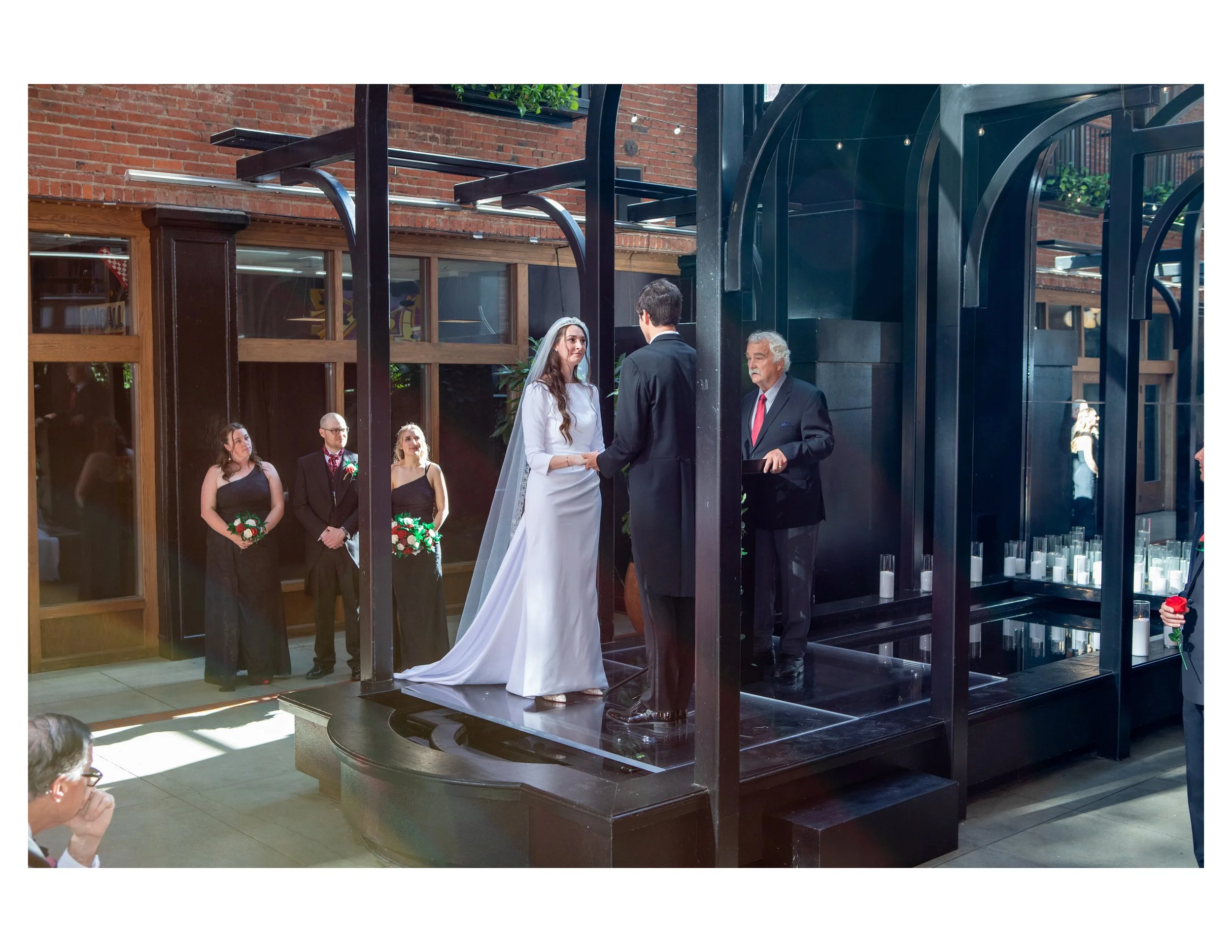 A wedding ceremony with a bride and groom standing on a black platform, exchanging vows, surrounded by several guests in formal attire, inside a venue with brick walls, large windows, and modern industrial decor.