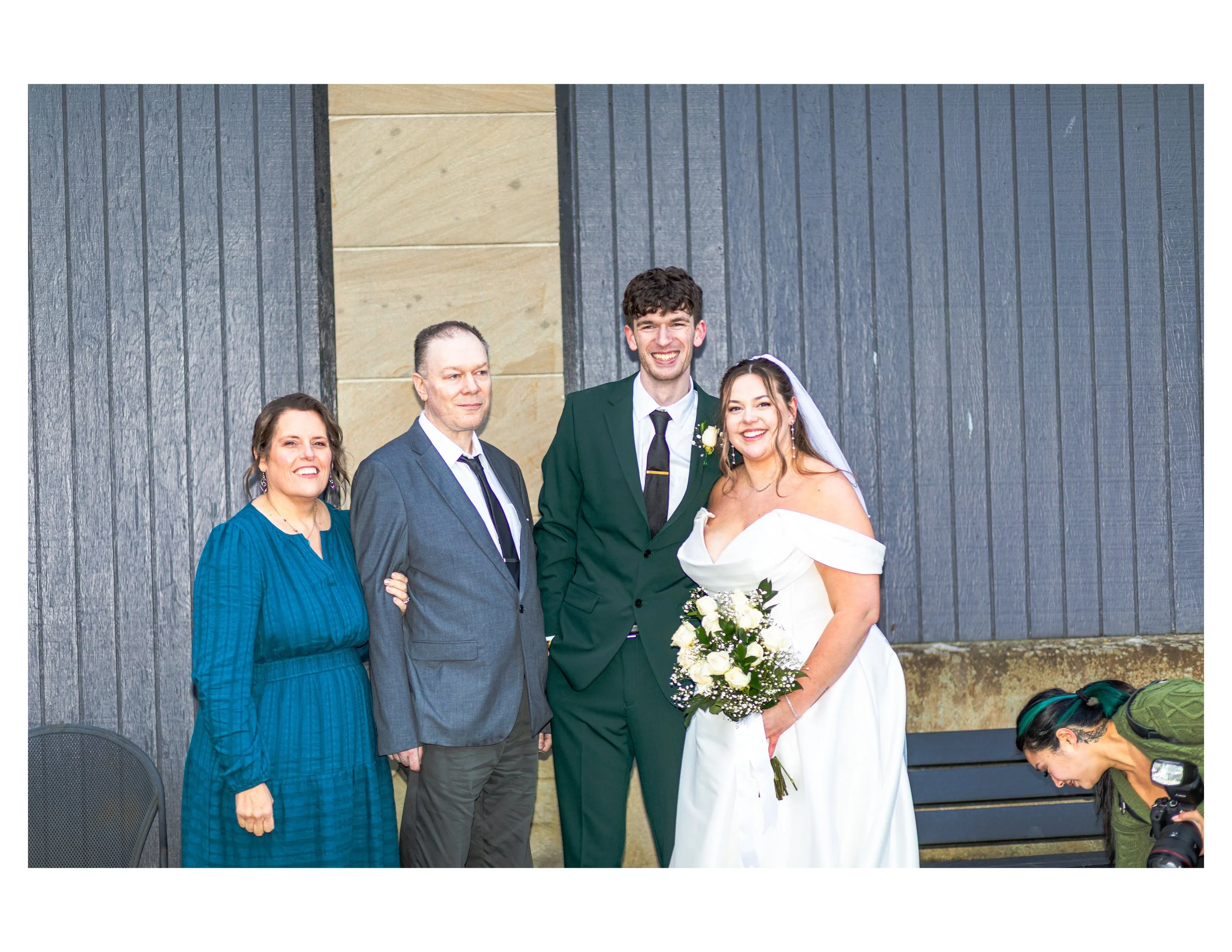 Group of five people at a formal event, including a woman in a blue dress, an older man in a gray suit, a smiling young man in a green suit, a woman in a wedding dress holding a bouquet, and a woman with colorful hair kneeling with a camera.