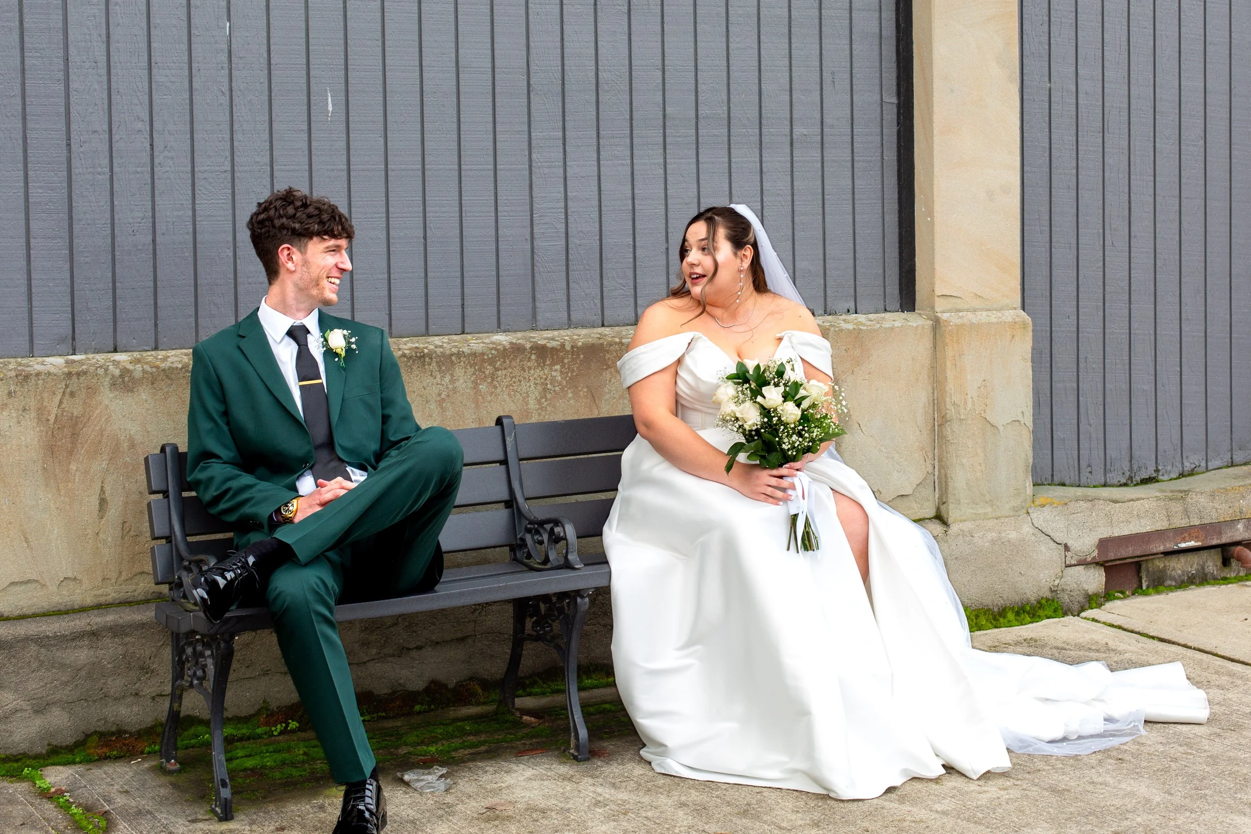 A bride and groom sitting on a bench outside, smiling and looking at each other during their wedding day. The bride is wearing a white wedding dress and holding a bouquet, while the groom is dressed in a green suit with a white shirt and black tie.