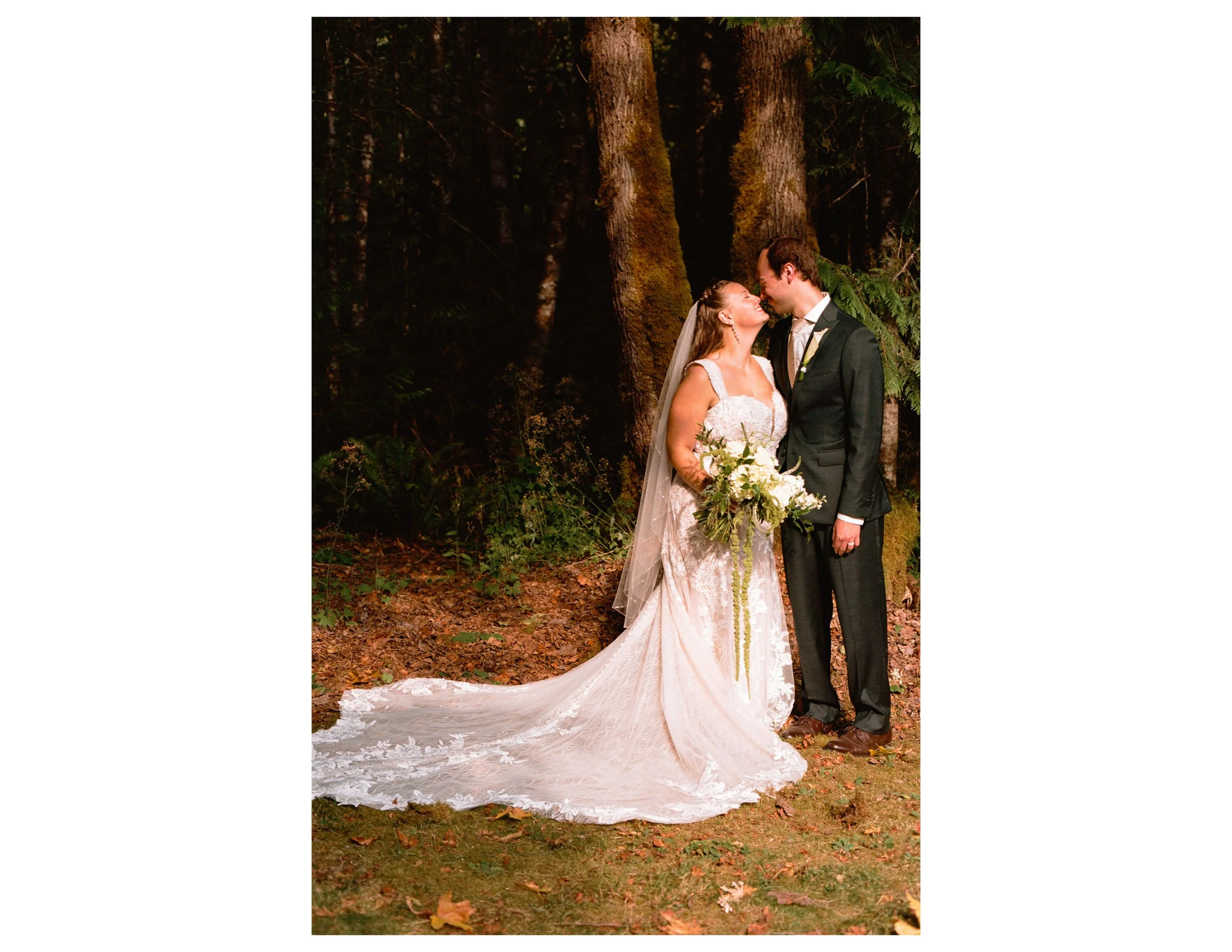 A bride and groom standing close together in a forest, smiling and looking into each other's eyes. The bride is wearing a white lace wedding gown with a long train and holding a bouquet, while the groom is in a dark suit with a boutonniere. The backg