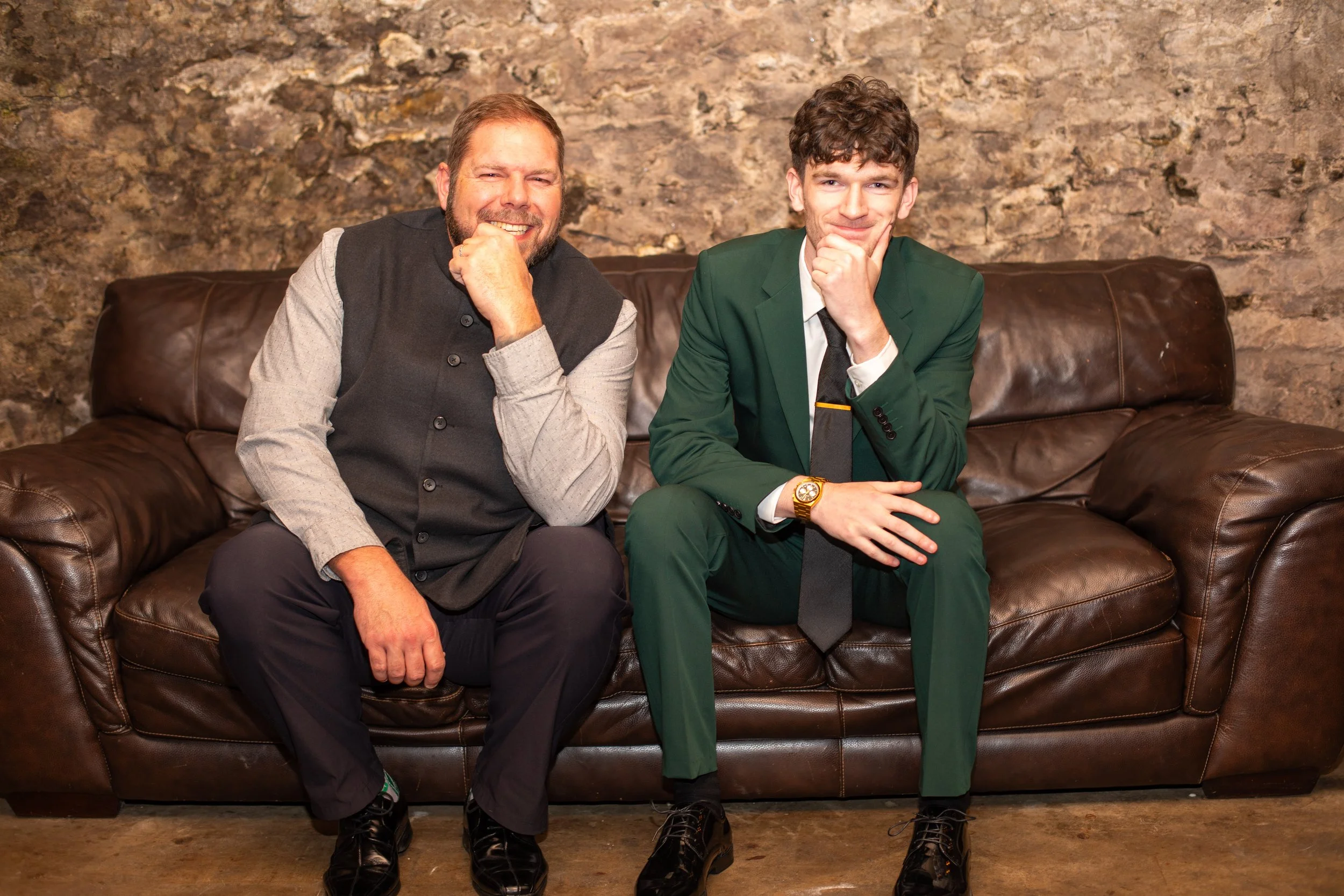 Two men sitting on a brown leather couch in front of a stone wall, smiling and posing thoughtfully with one hand on their chin.
