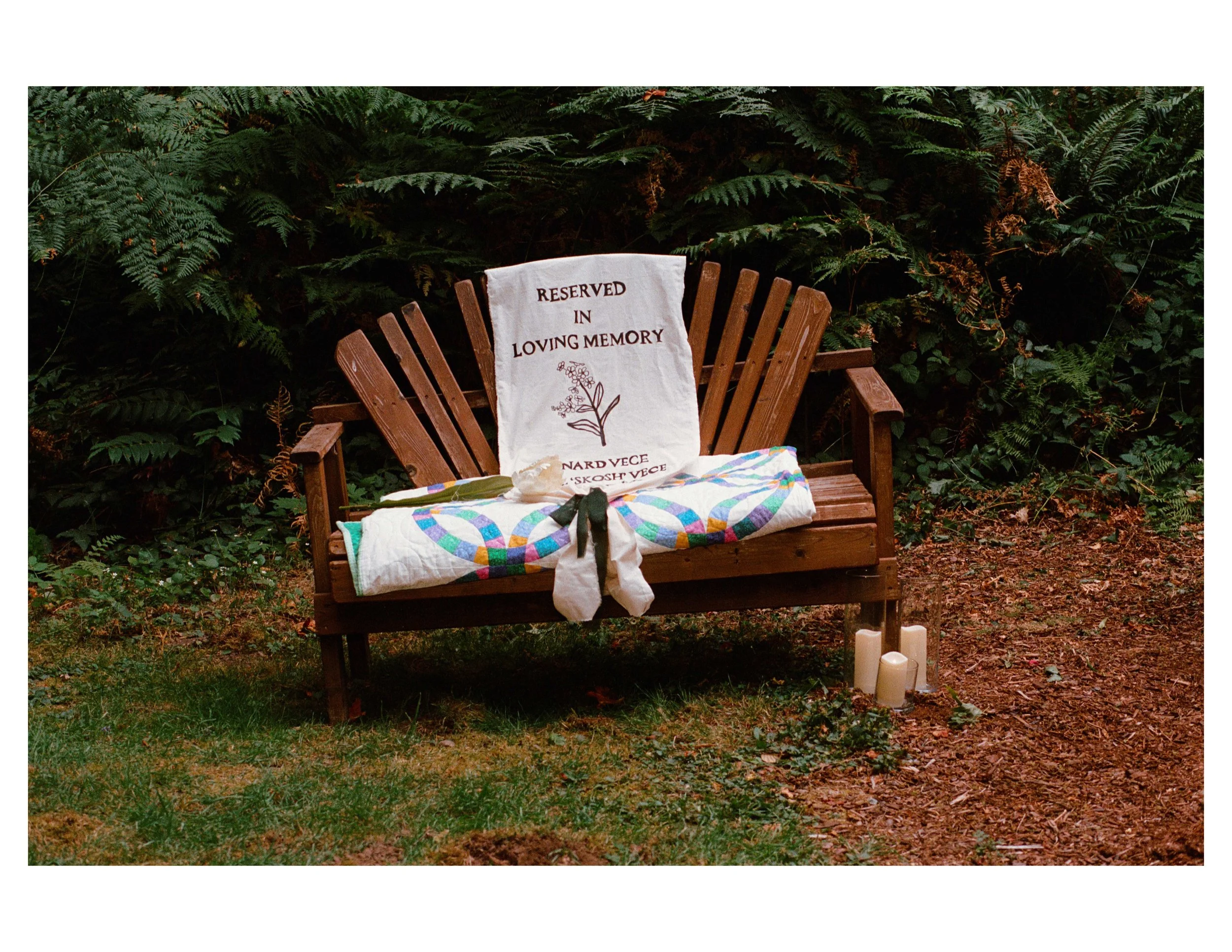 A wooden bench outdoors surrounded by green ferns and plants, with a white cloth draped over the backrest that reads 'Reserved in Loving Memory' and has a floral illustration, and candles placed on the ground nearby.