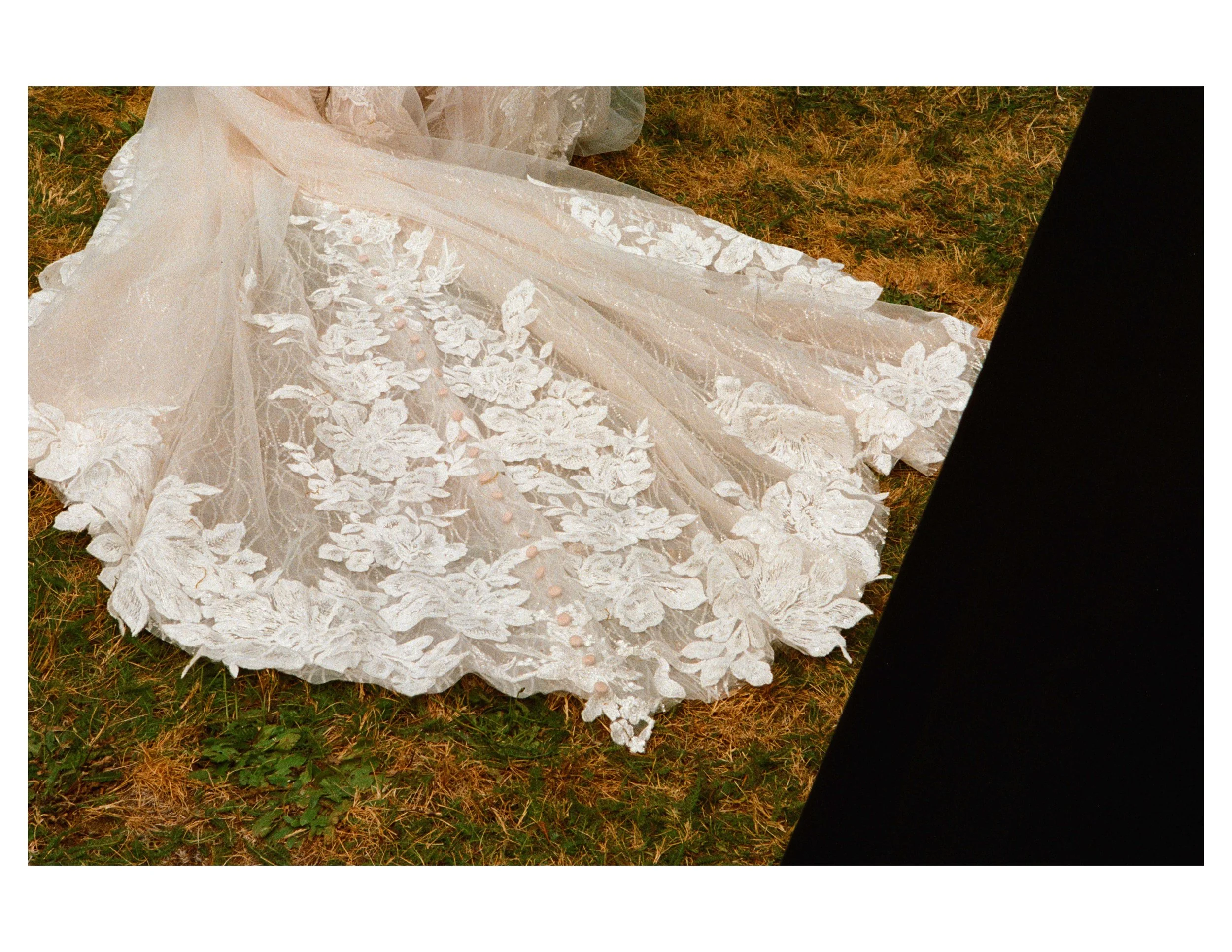 A white wedding dress with lace embroidery rests on grass, partially covered by a black fabric.