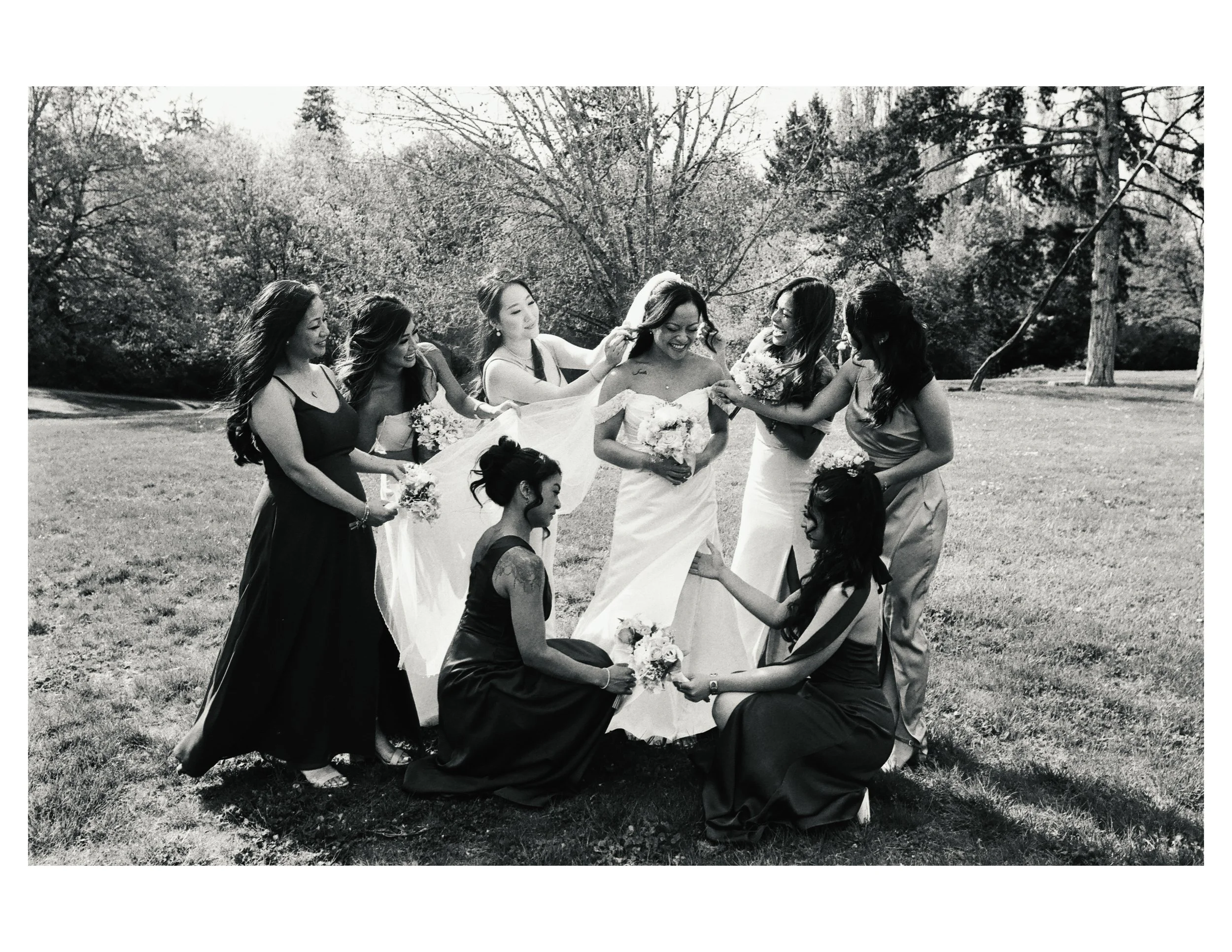 Group of women in bridesmaid dresses surrounding a bride in a wedding dress outdoors, with trees in the background, some women holding bouquets, in black and white.
